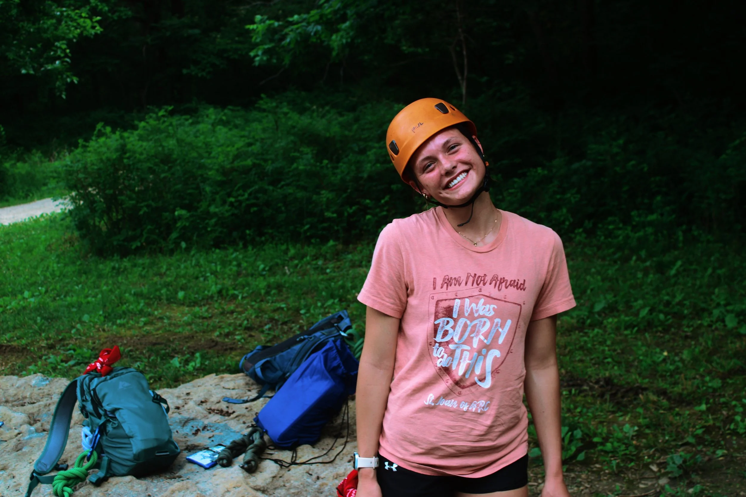 Smiling woman wearing an orange helmet and pink t-shirt standing outdoors in a wooded area with backpacks and climbing gear on the ground nearby.