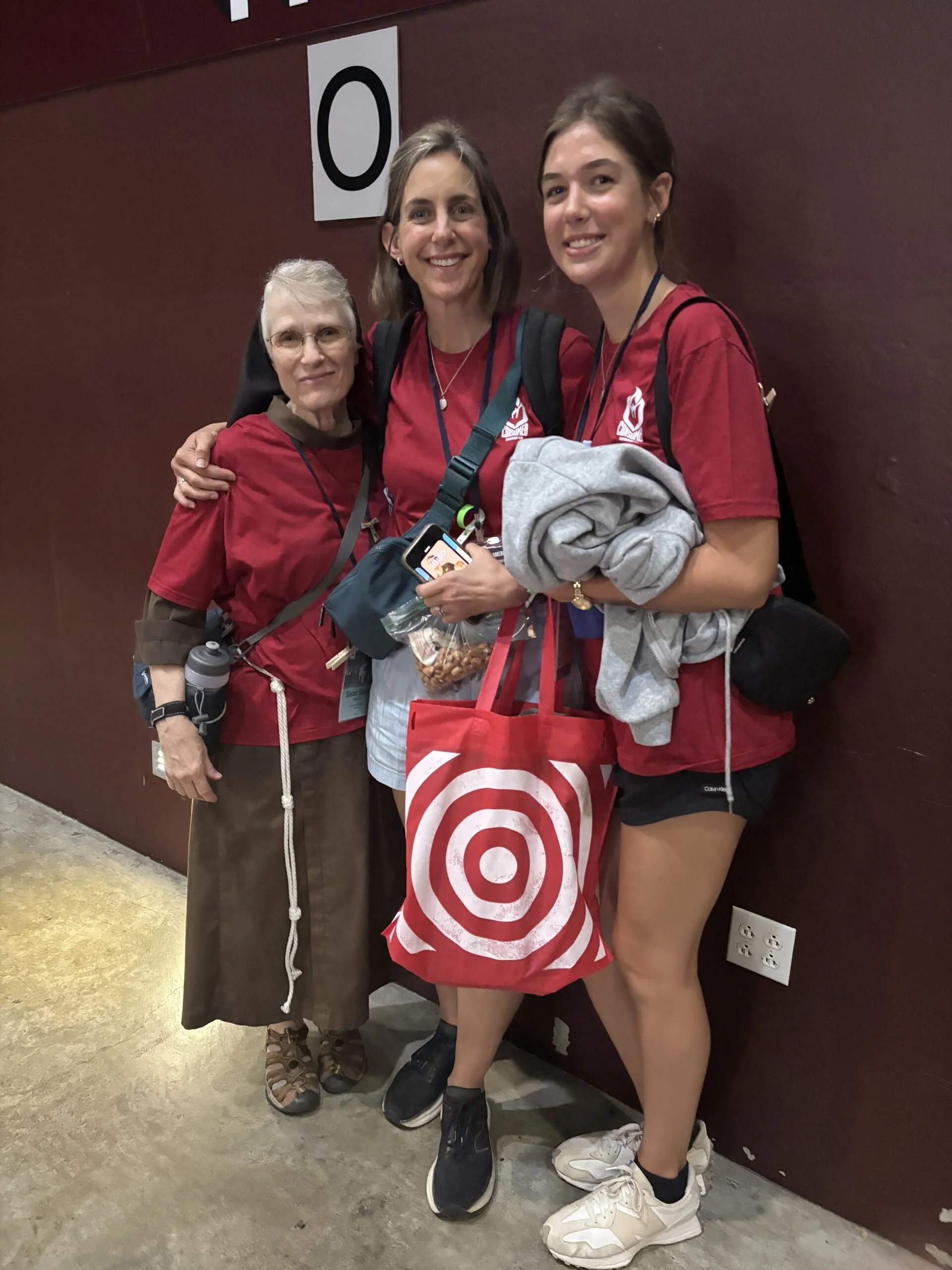 Three women posing together, wearing red shirts with a white logo, standing in front of a maroon wall with a large number 0.