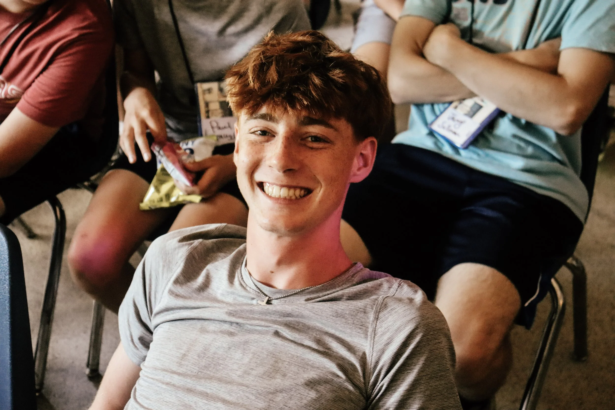 Smiling young man taking a selfie at a casual gathering, surrounded by others, seated in chairs.