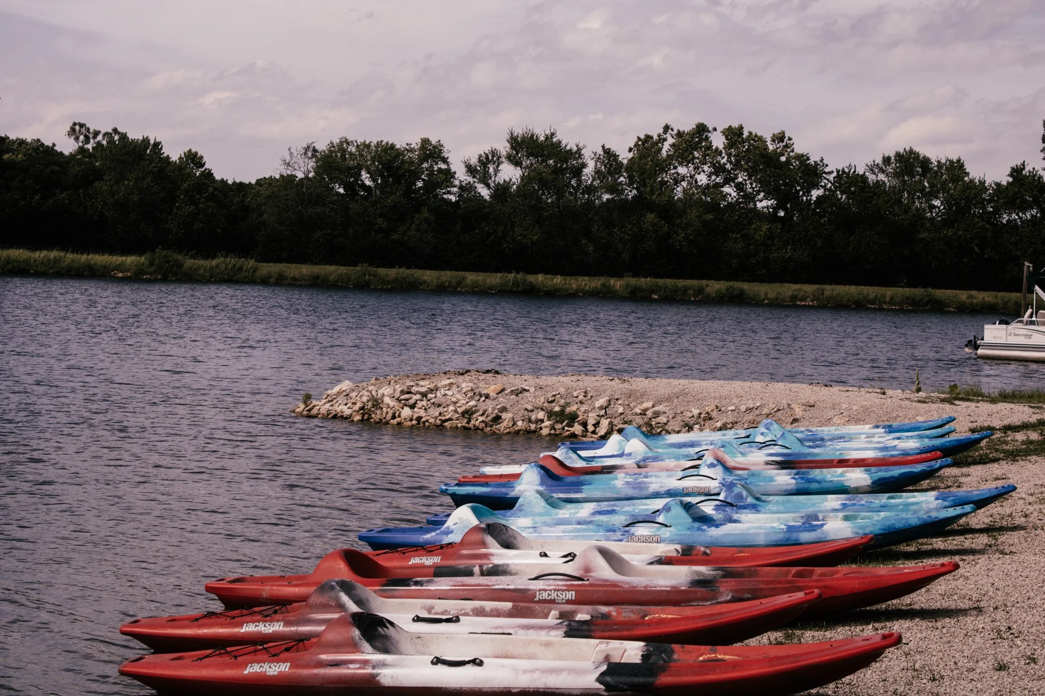 Colorful kayak boats lined up on the shore of a body of water with trees and cloudy sky in the background.