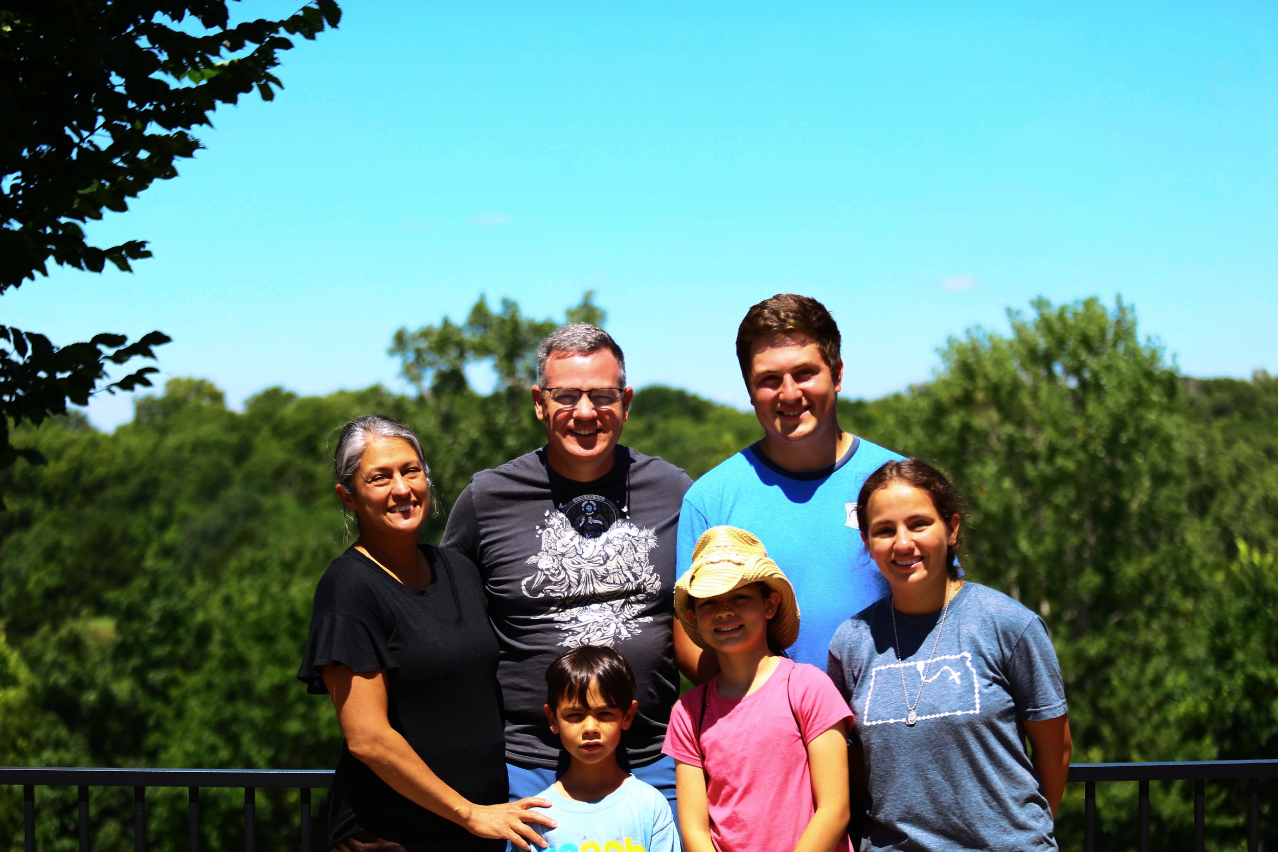 A group of seven people, including children and adults, standing outdoors on a sunny day with green trees in the background. They are smiling and looking at the camera. Some are wearing casual clothing and hats.
