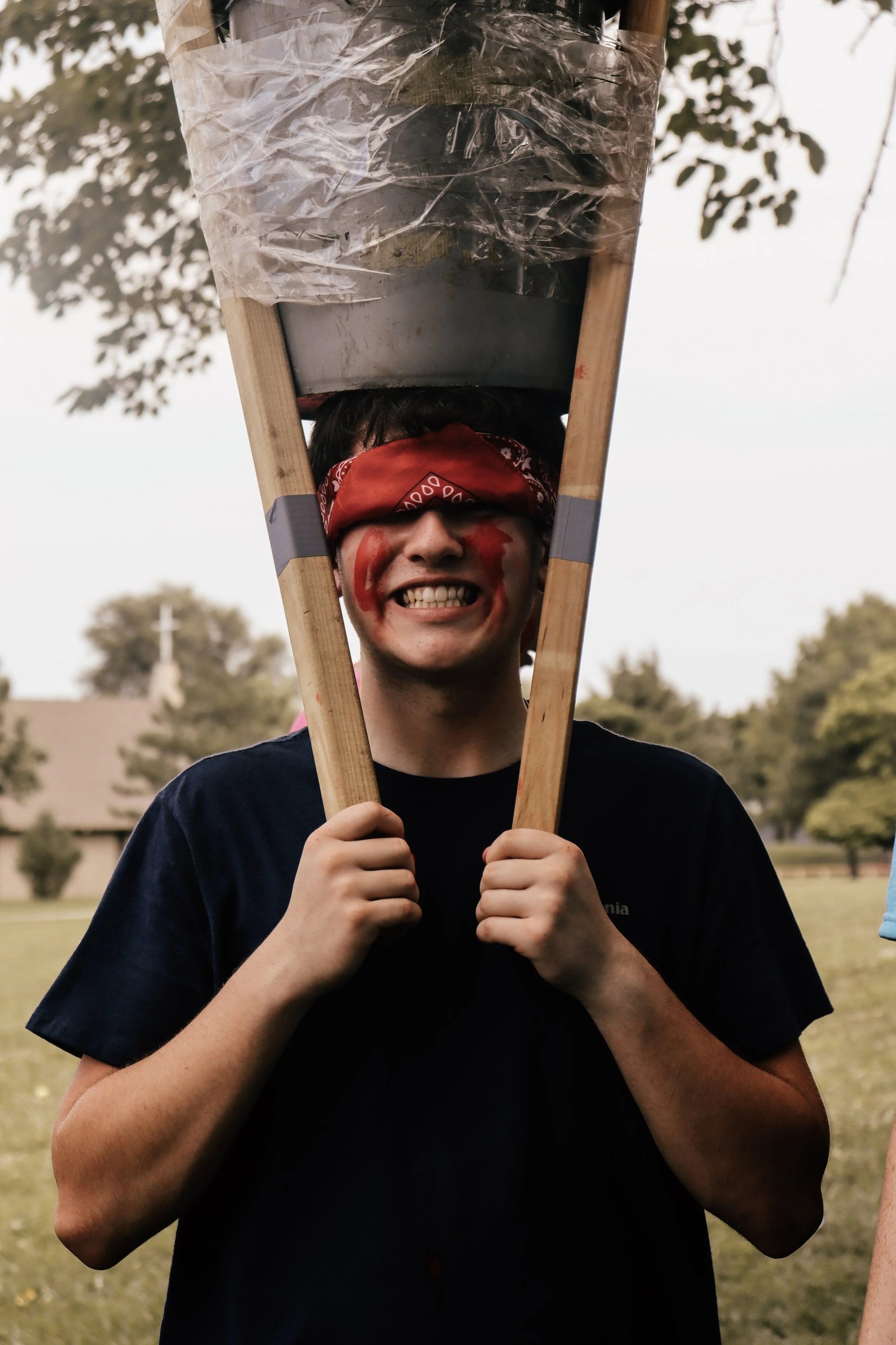 A young man with paint on his face and a red bandana covering his eyes, smiling, holding a wooden ladder with a plastic bag on top.