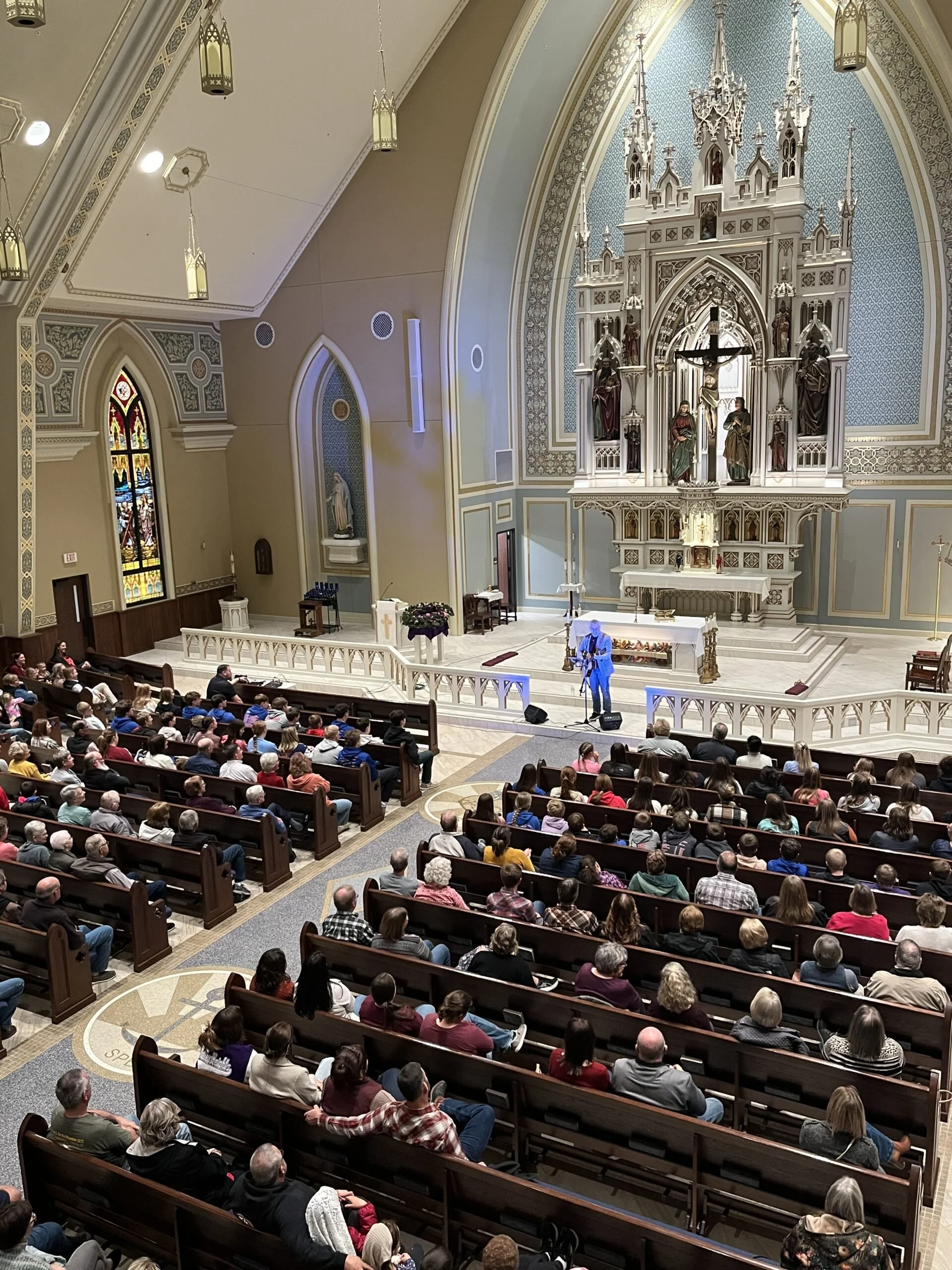 Inside a church with a congregation seated in wooden pews, a man dressed in blue stands at a microphone near the altar, which has a large elaborate white and gold altar with religious statues and a crucifix. The church features stained glass windows 