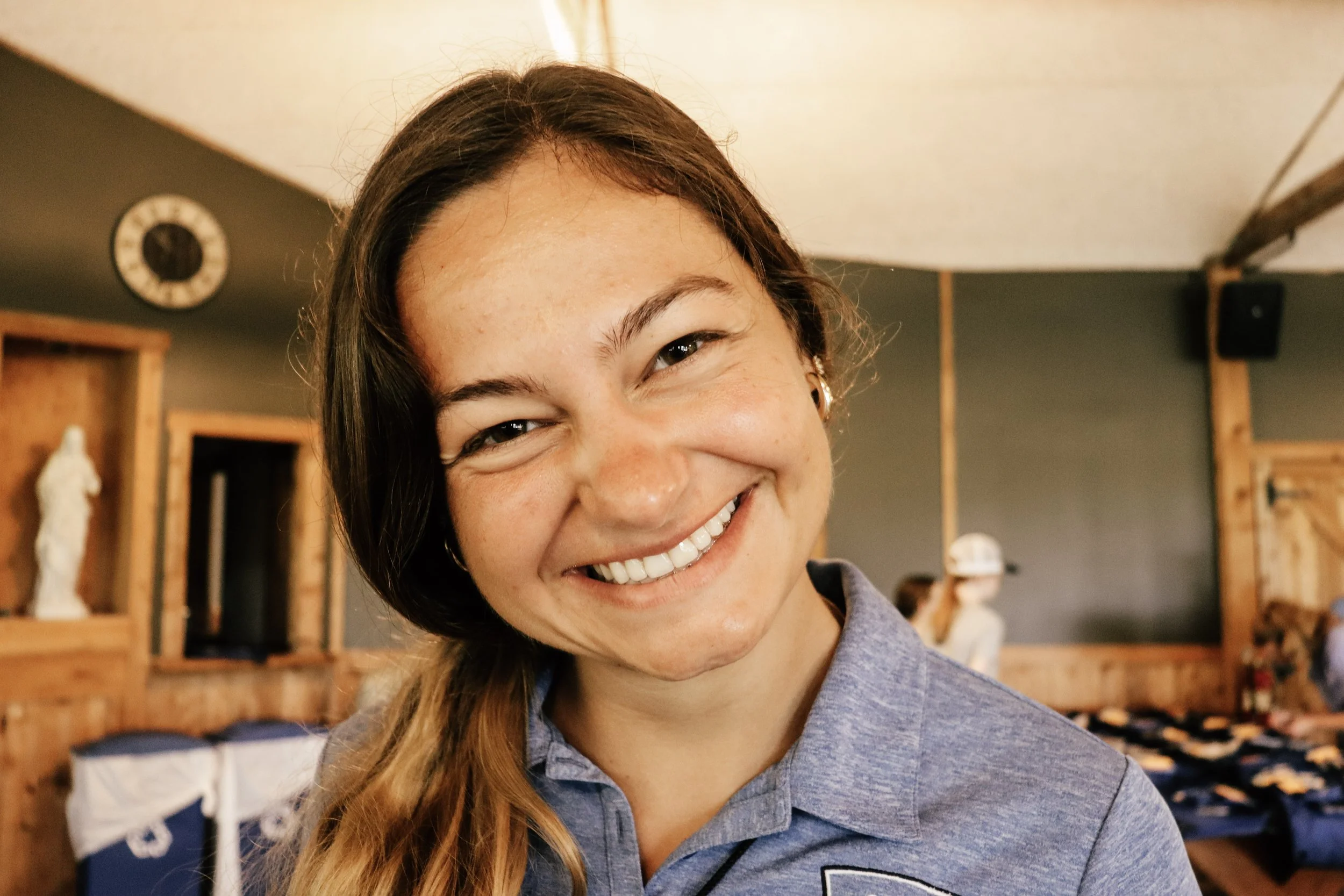 A smiling young woman with dark hair and hoop earrings takes a selfie in a cozy room with wooden shelving and art pieces in the background.