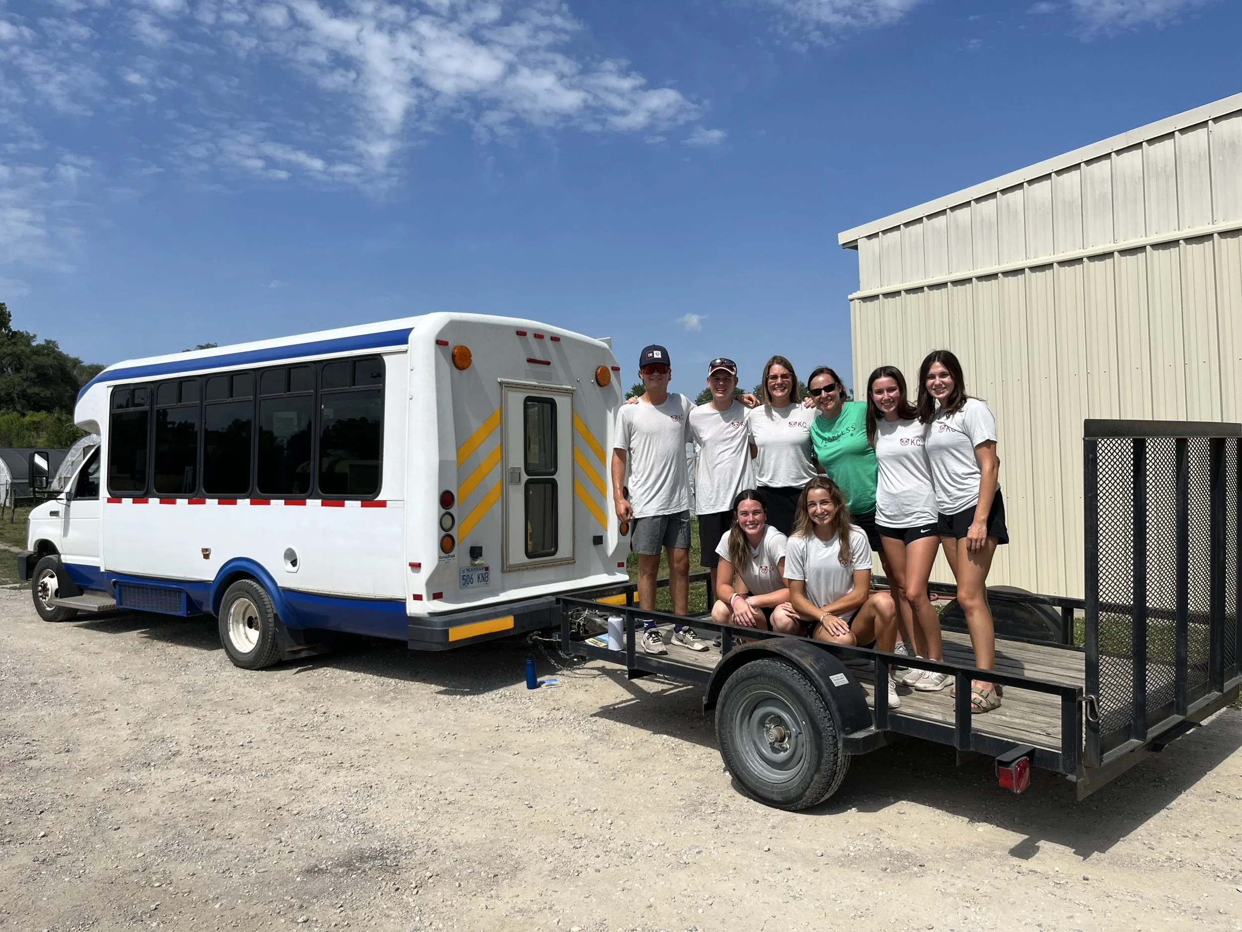 Group of six young women and two young men posing together outdoors next to a white and blue bus and a black trailer with a metal gate, under a partly cloudy sky.