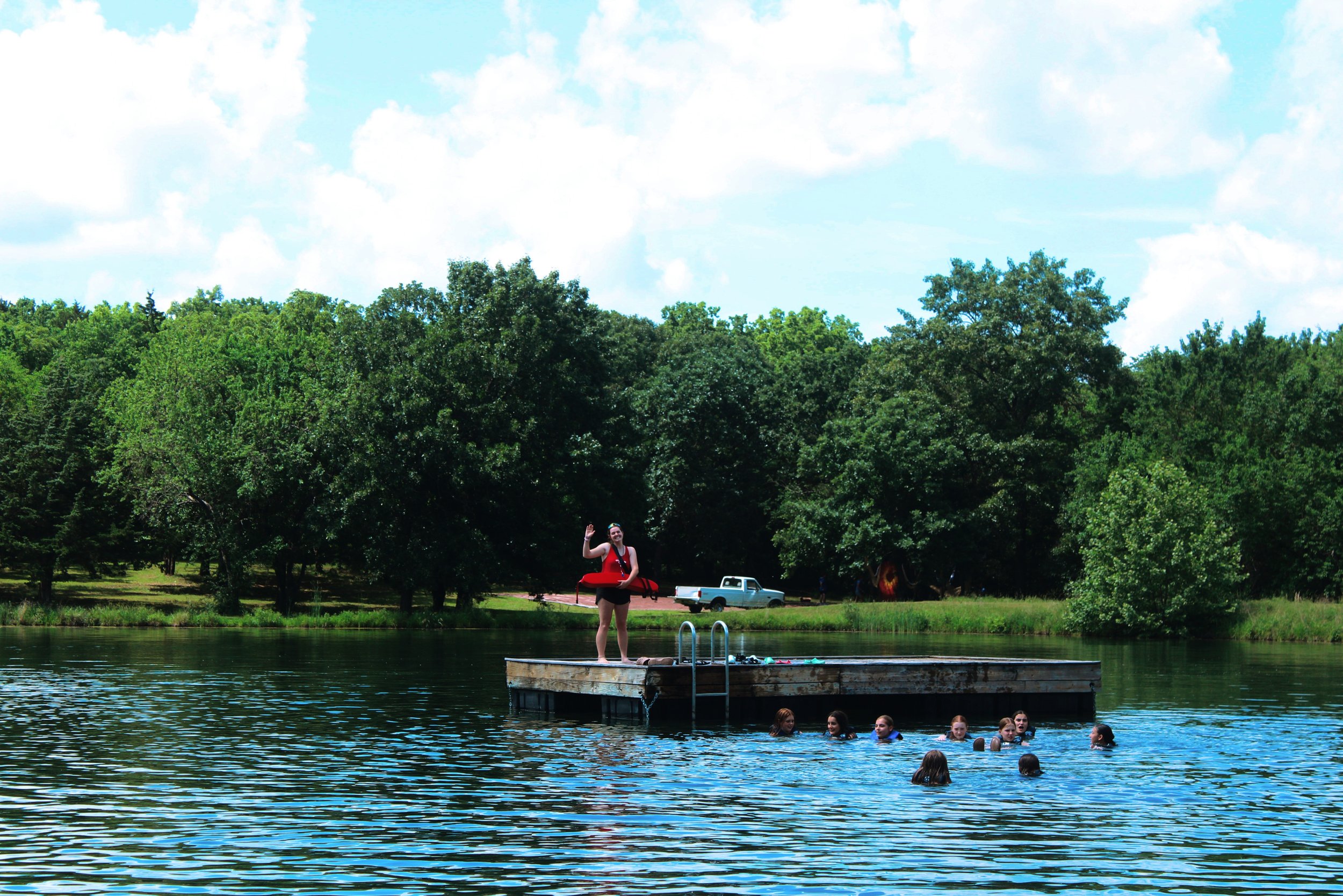 People enjoying swimming and floating on a lake with a dock, surrounded by green trees and lush foliage under a partly cloudy sky.