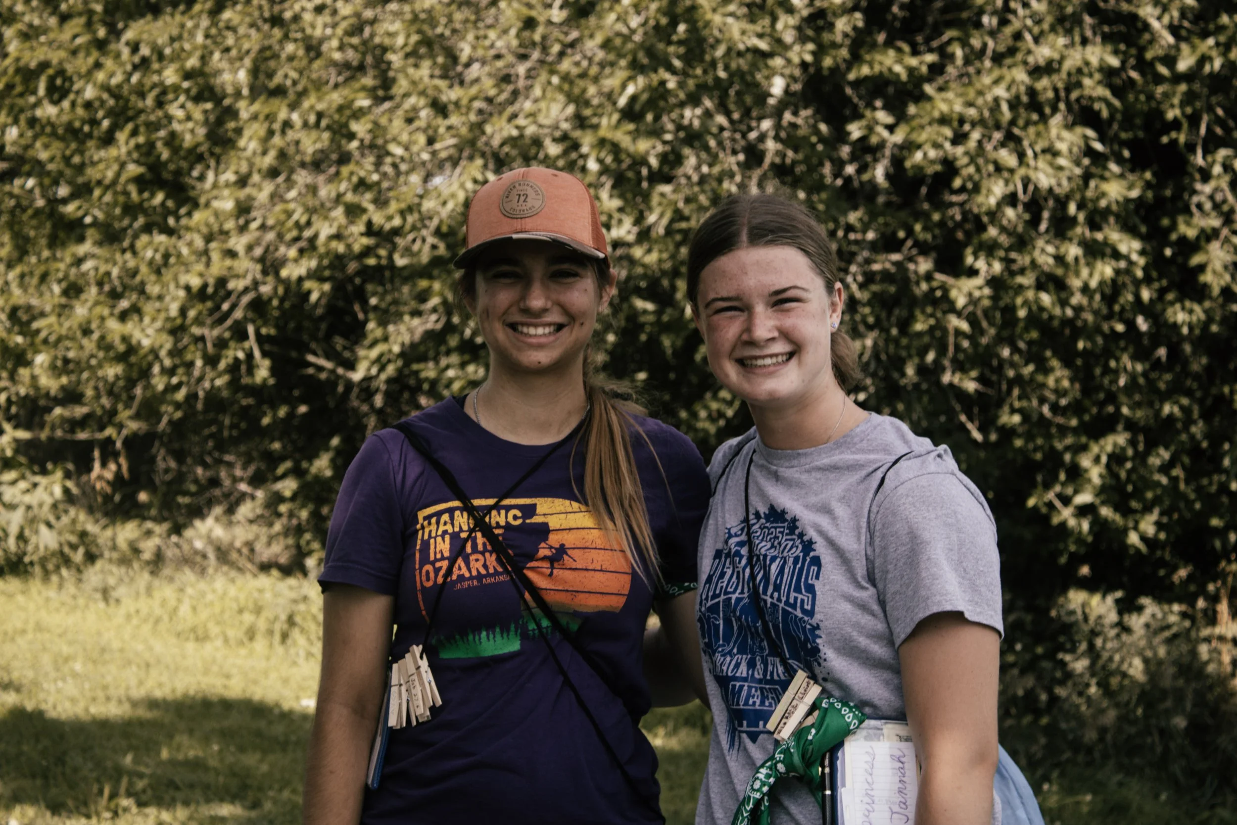 Two young women standing outdoors in front of green foliage, smiling at the camera, wearing casual t-shirts with bandanas tied around their waists.