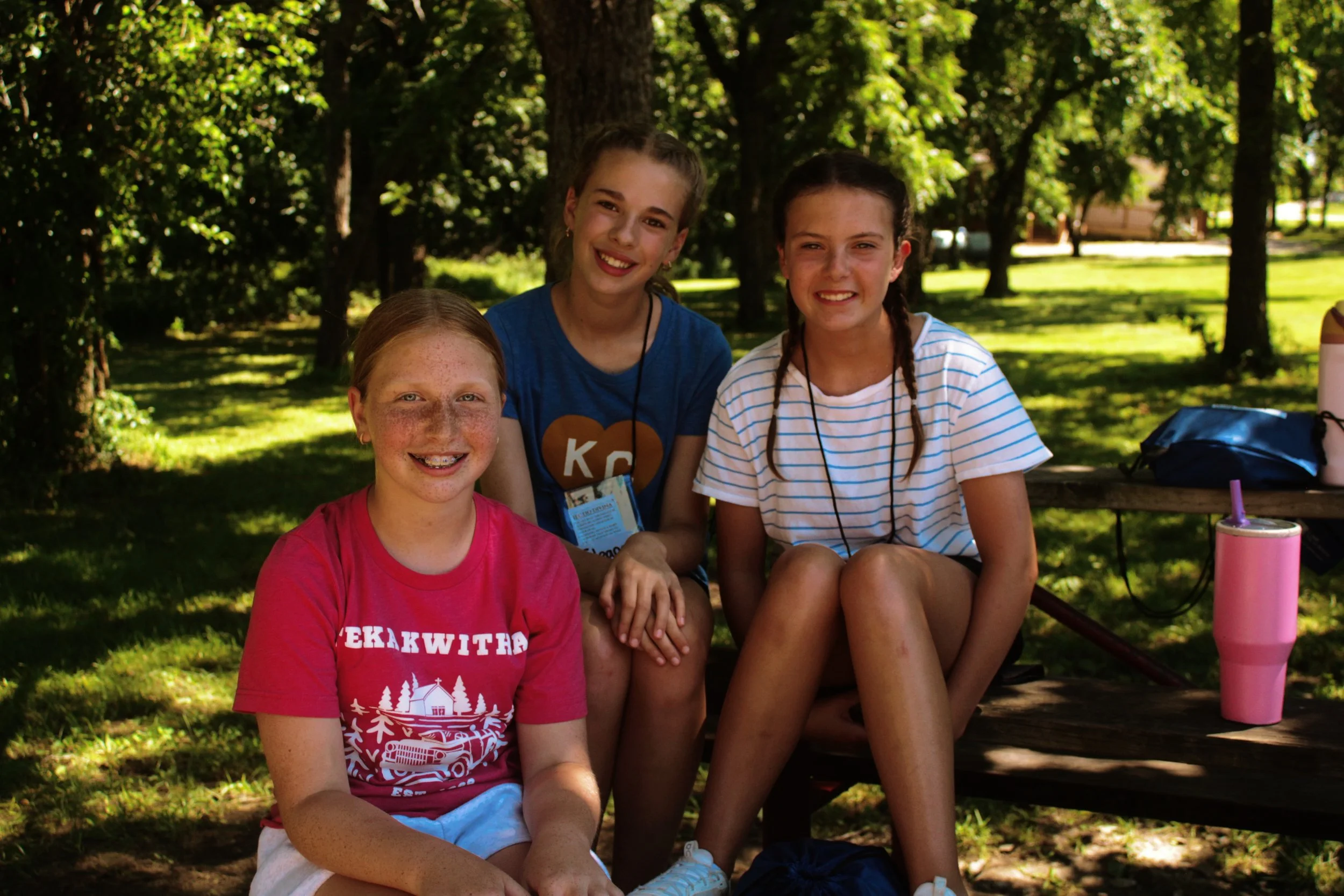 Four young girls sitting outdoors in a shaded park, smiling at the camera. They are surrounded by green trees and grass, with some camping items on a bench beside them.