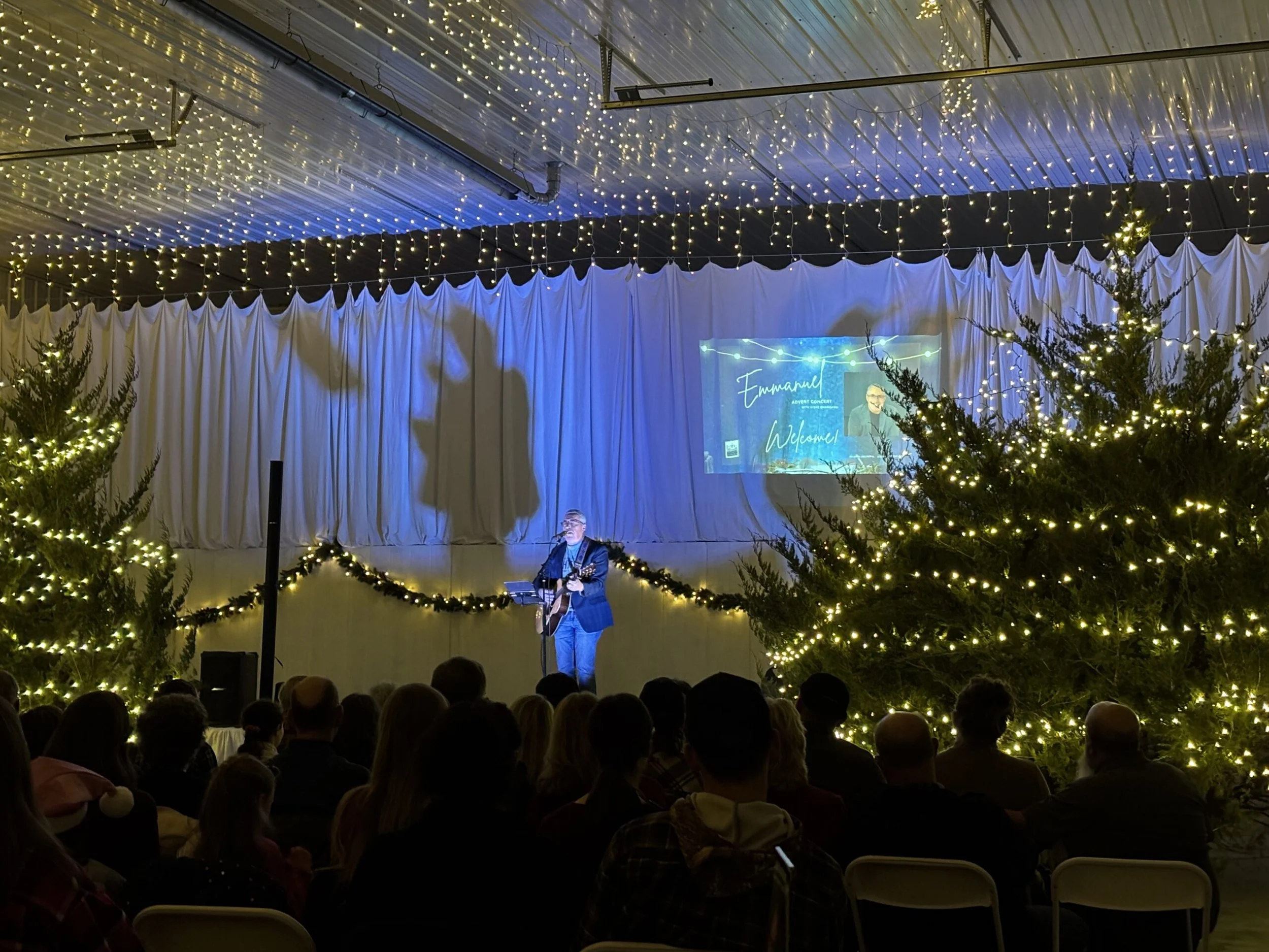 Indoor concert scene with Christmas trees decorated with lights framing the stage. Stage has white drapes and a large screen projecting a Facebook event for Emanuel Advent Concert, featuring a male singer playing guitar. The ceiling is decorated with