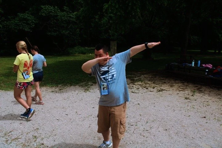 Young man dabbing outdoors at a park with other people in the background.