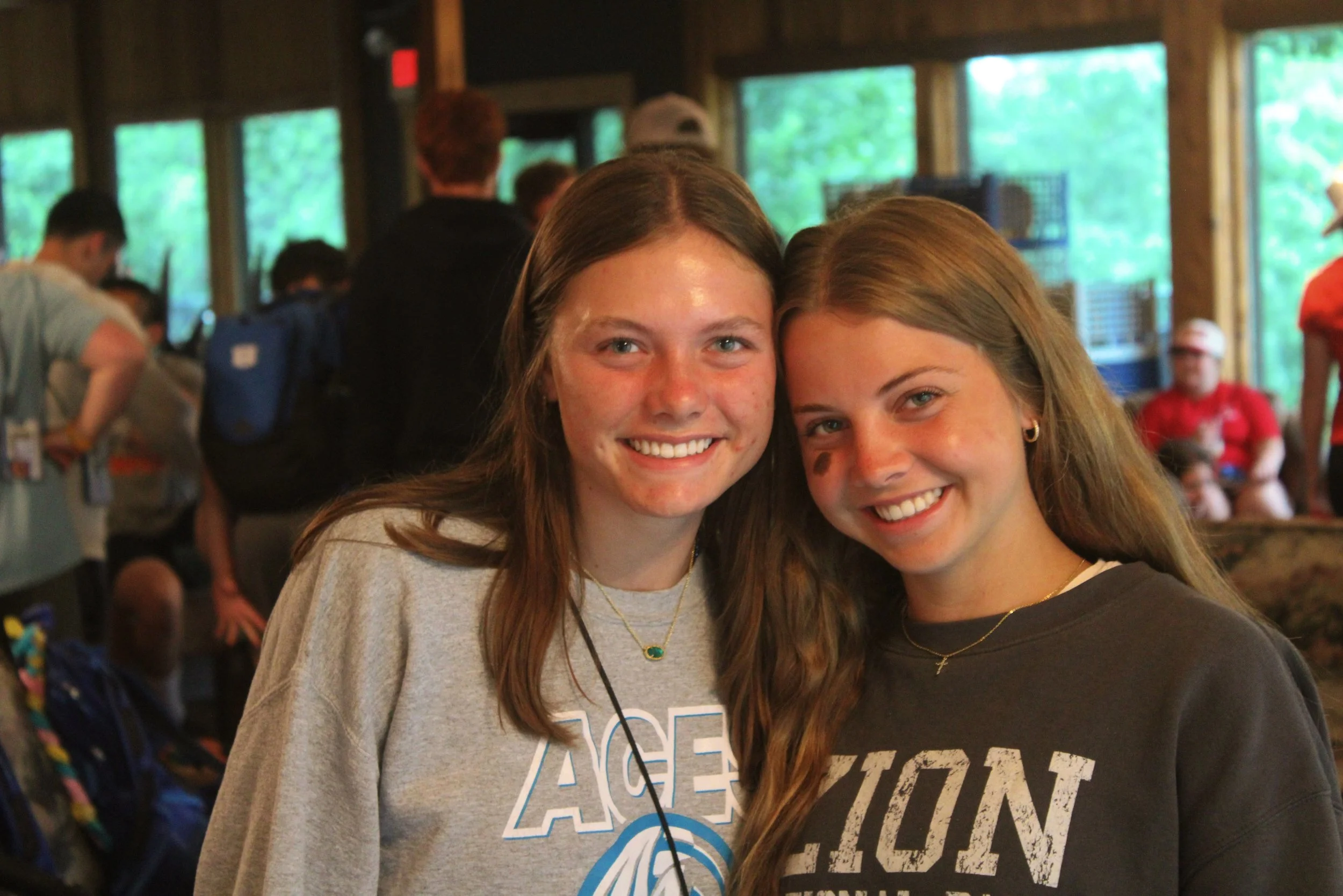 Two young women are smiling and posing for a photo indoors, with a group of people in the background engaged in conversation or activities.
