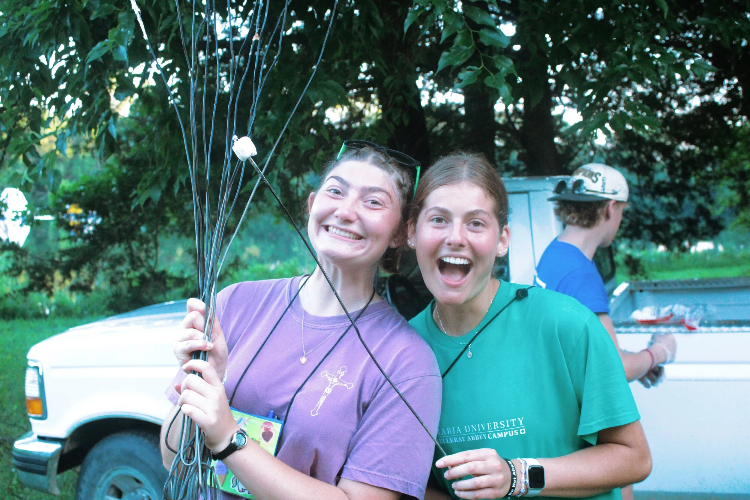 Two young women smiling at the camera, holding some wires or cables, standing outdoors with trees in the background. A young man is in the background, facing away, wearing a hat, near a vehicle.