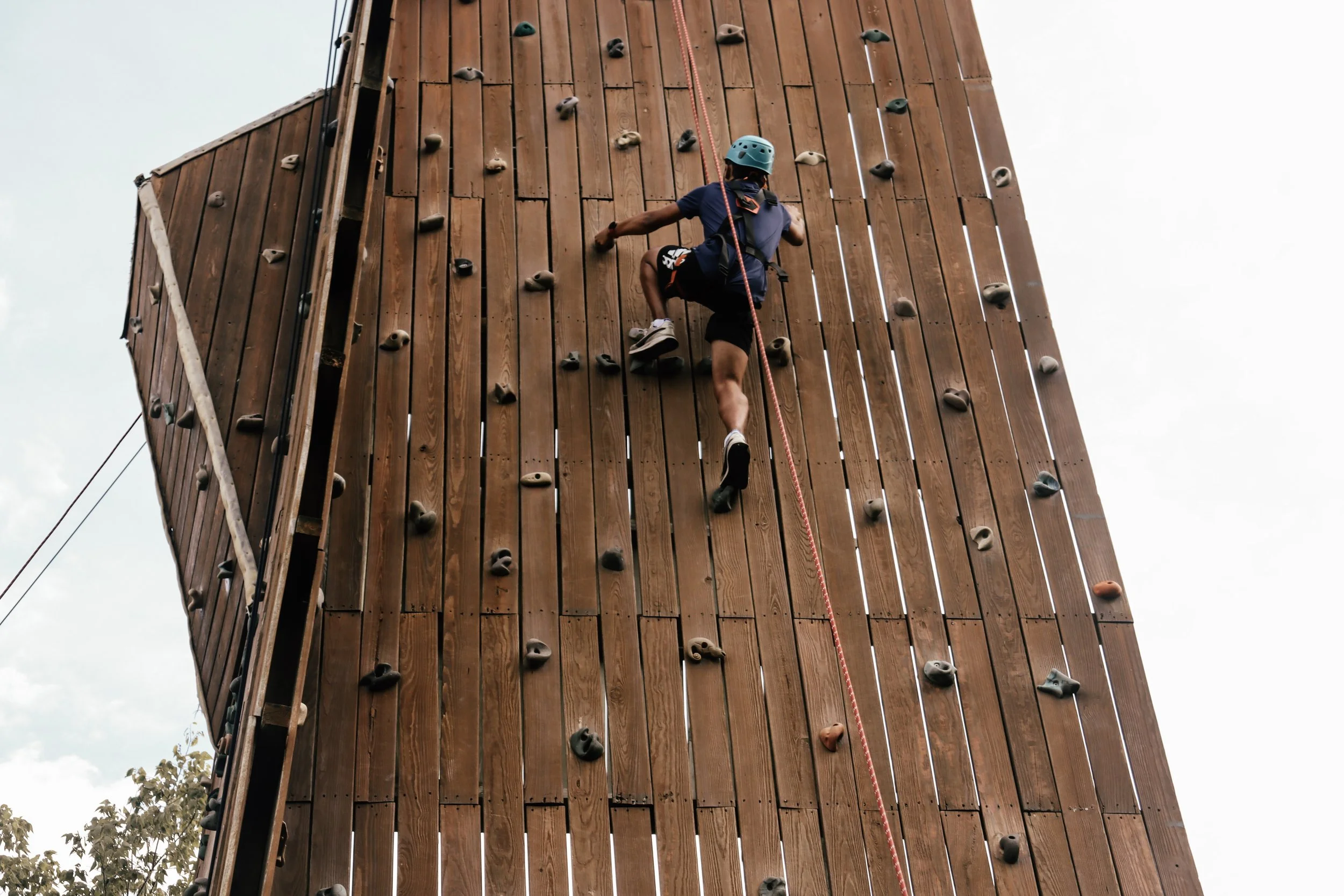 A person wearing a helmet and harness climbing an outdoor wooden rock wall with various handholds and footholds.