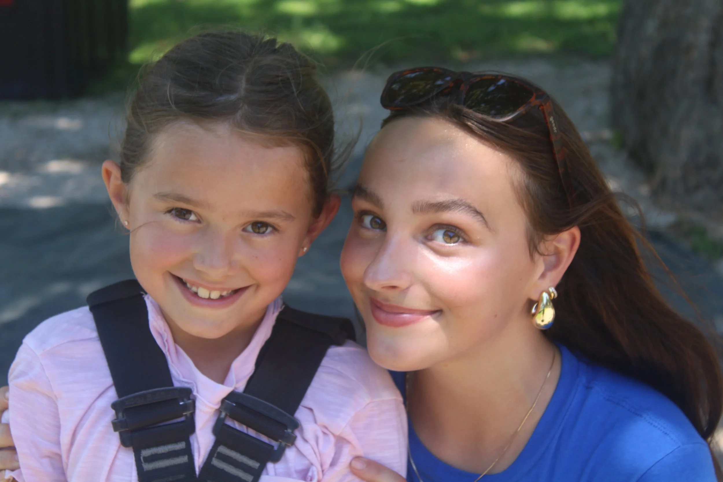 A young girl and a woman smiling outdoors with green trees in the background.