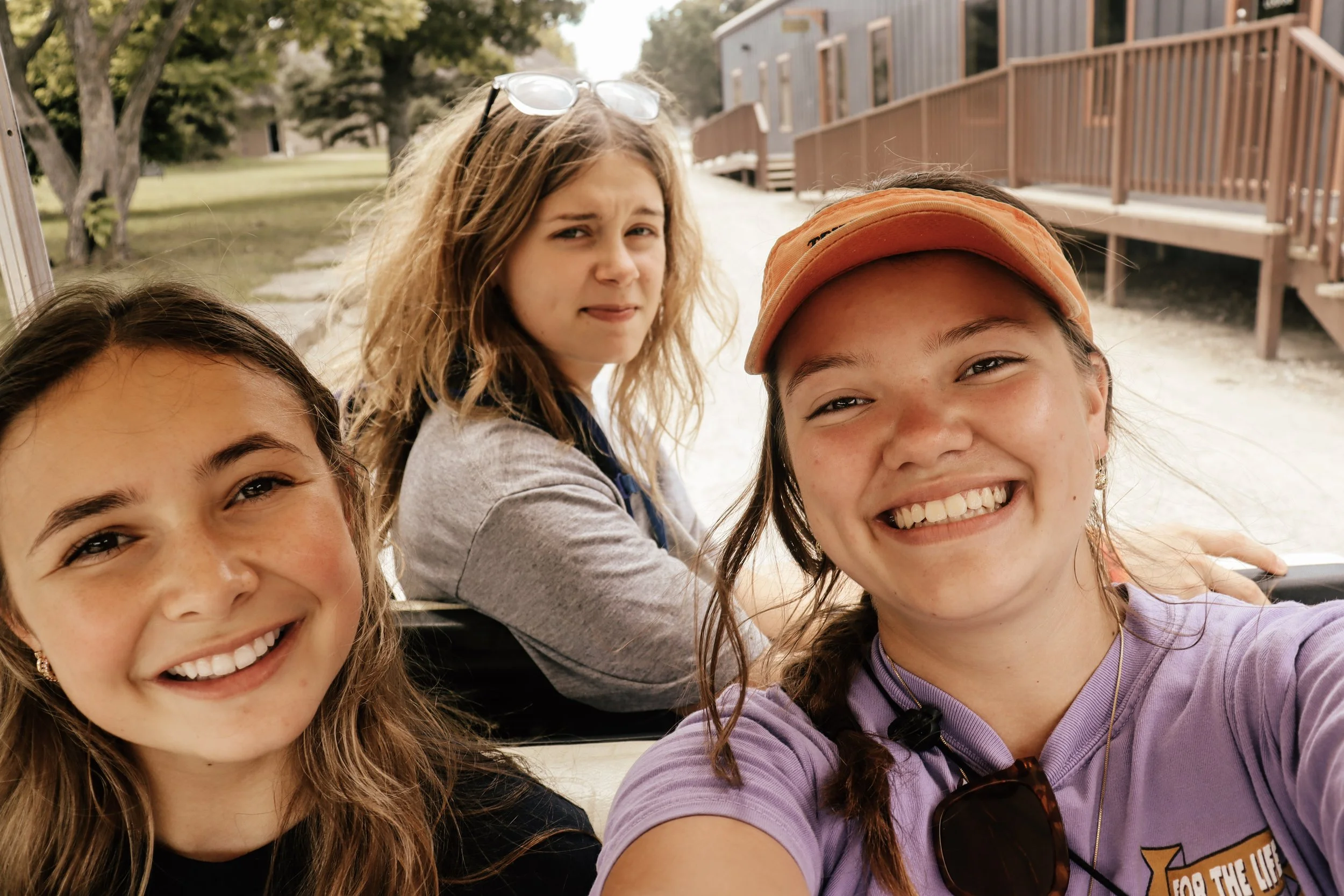 Four young girls smiling and taking a selfie outdoors near a wooden building and trees.