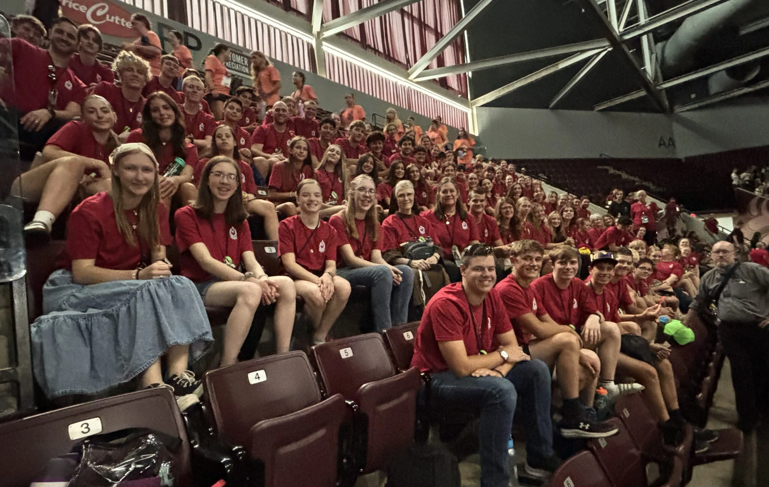 Group of young people wearing red shirts sitting in stadium seats at an indoor sports arena.