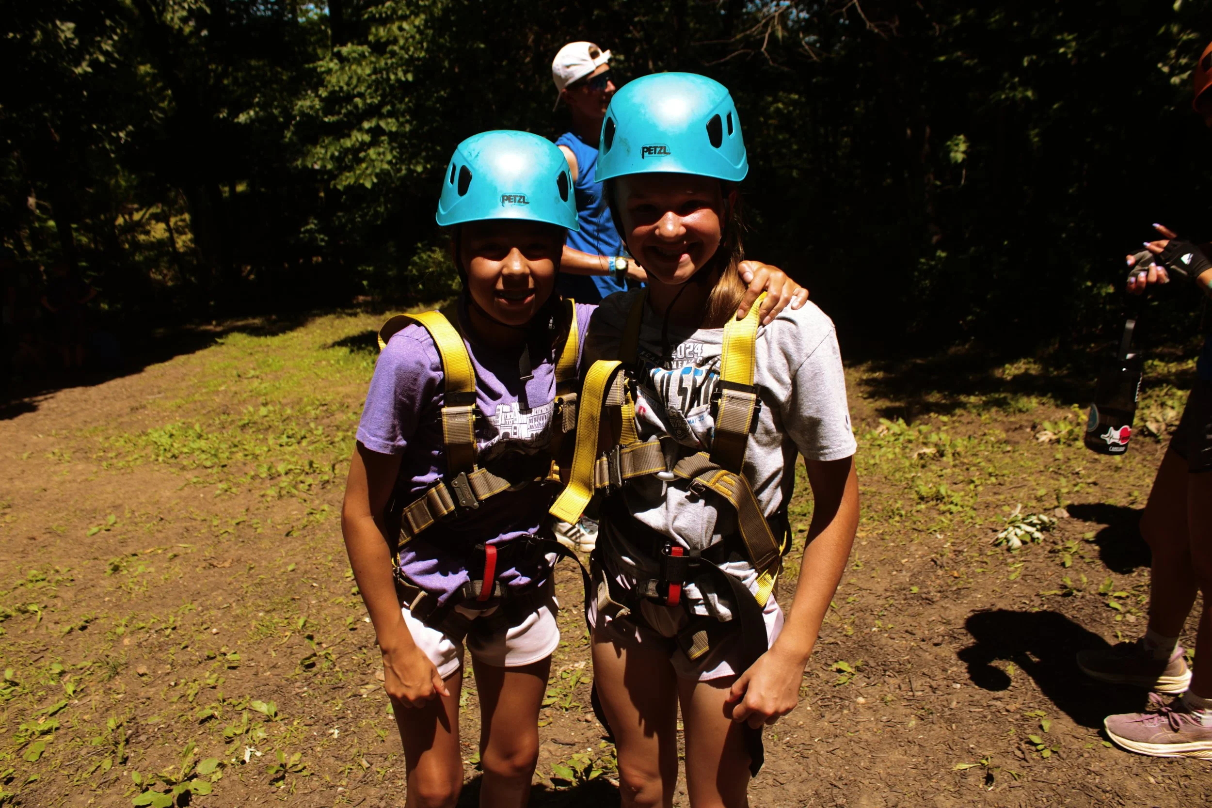 Two girls wearing blue helmets and harnesses stand outdoors, smiling and posing for a photo with trees in the background.