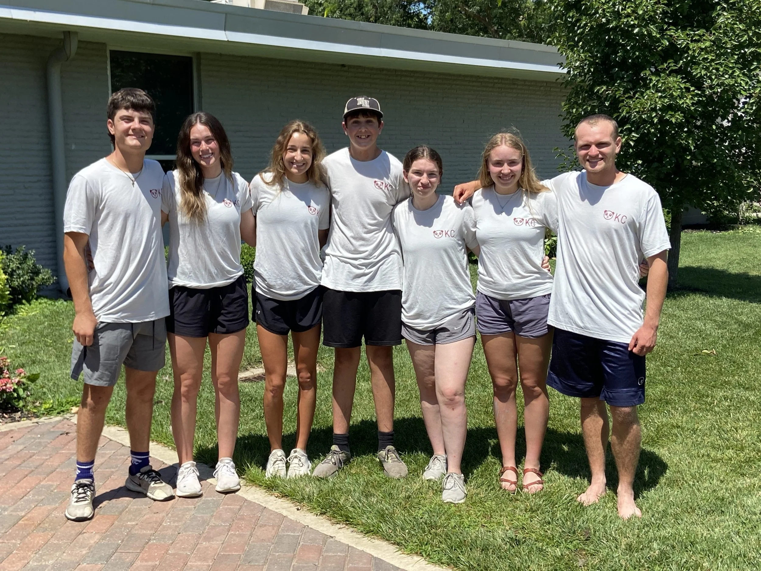 A group of seven young people standing outside on a sunny day, smiling at the camera. They are wearing white T-shirts with a pink pig logo and the letters 'KC' printed on them. The background includes a house with white siding, a window, green bushes, and trees.