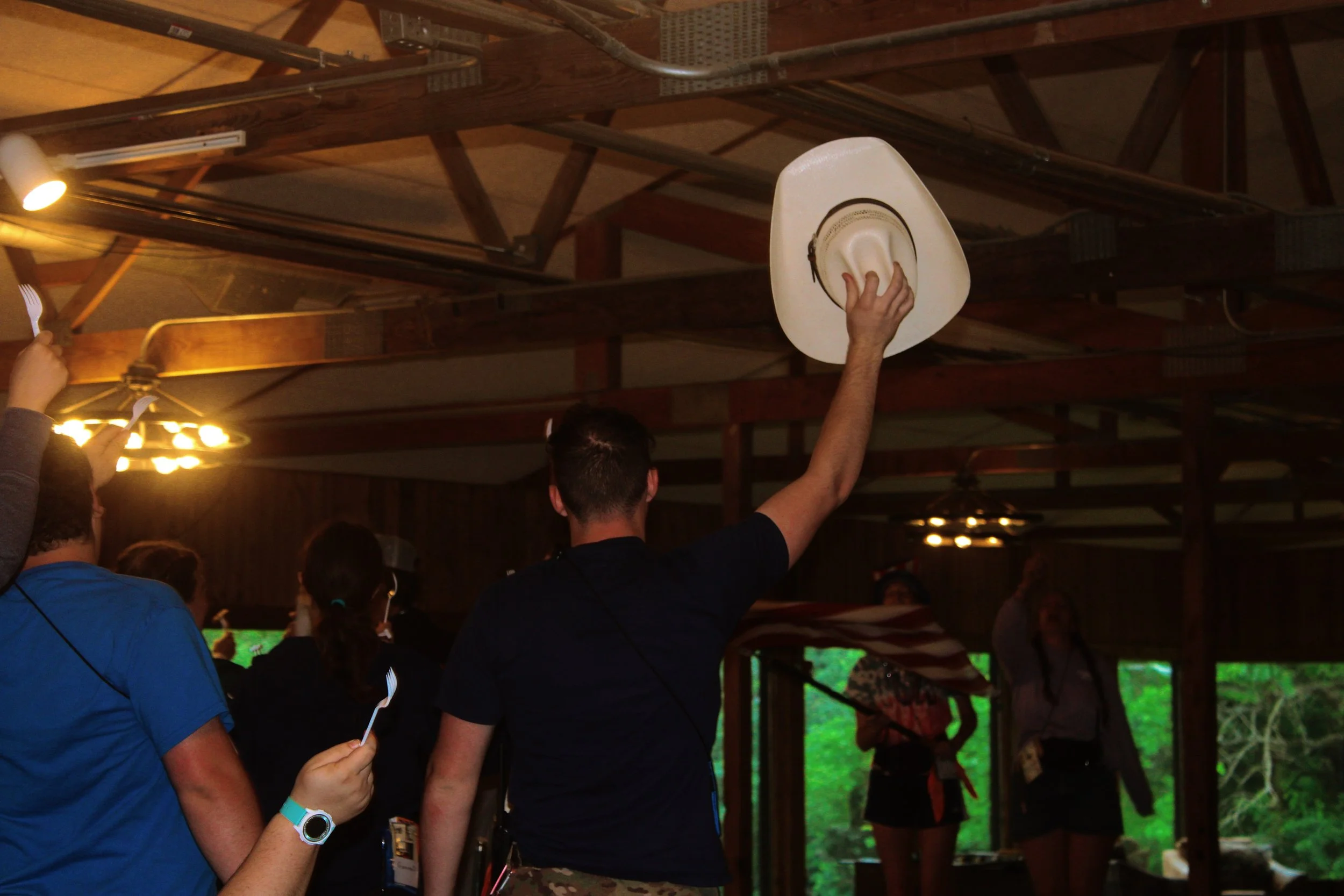 People indoors, with one person holding a white cowboy hat with black accents and a brown band, and others holding paper fans. The ceiling has exposed wooden beams and lighting fixtures, with a green outdoor scene visible through a window in the back