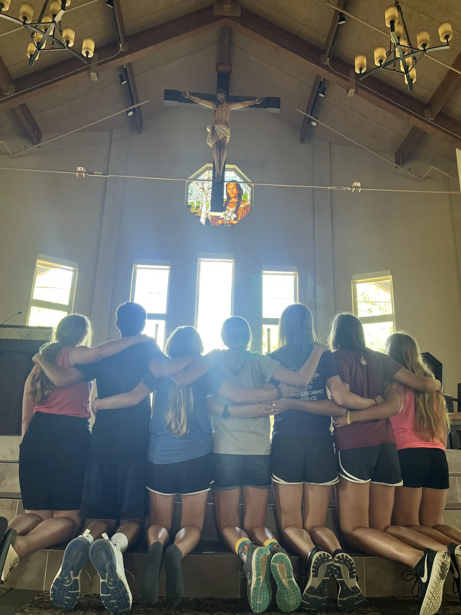 A group of young women sitting on a bench with their backs to the camera, arms around each other's shoulders, inside a church with a crucifix and stained glass window behind them.