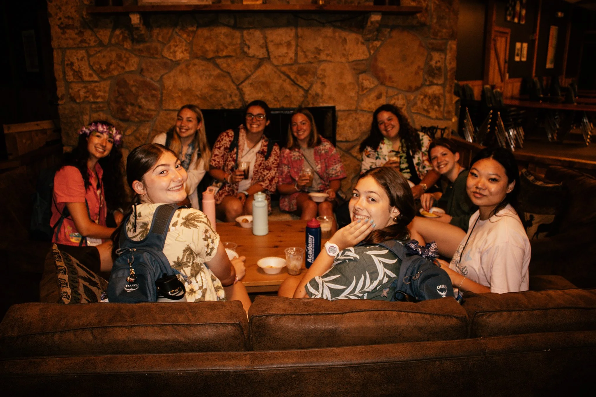 Group of eight young women smiling and sitting around a wooden table in a cozy restaurant with a stone fireplace in the background. Some hold drinks, and one wears a flower crown.