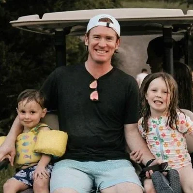 Three people sitting on a golf cart: a man in a backwards cap and black shirt, a young girl with a yellow life jacket, and a young girl with a floral dress, all smiling.