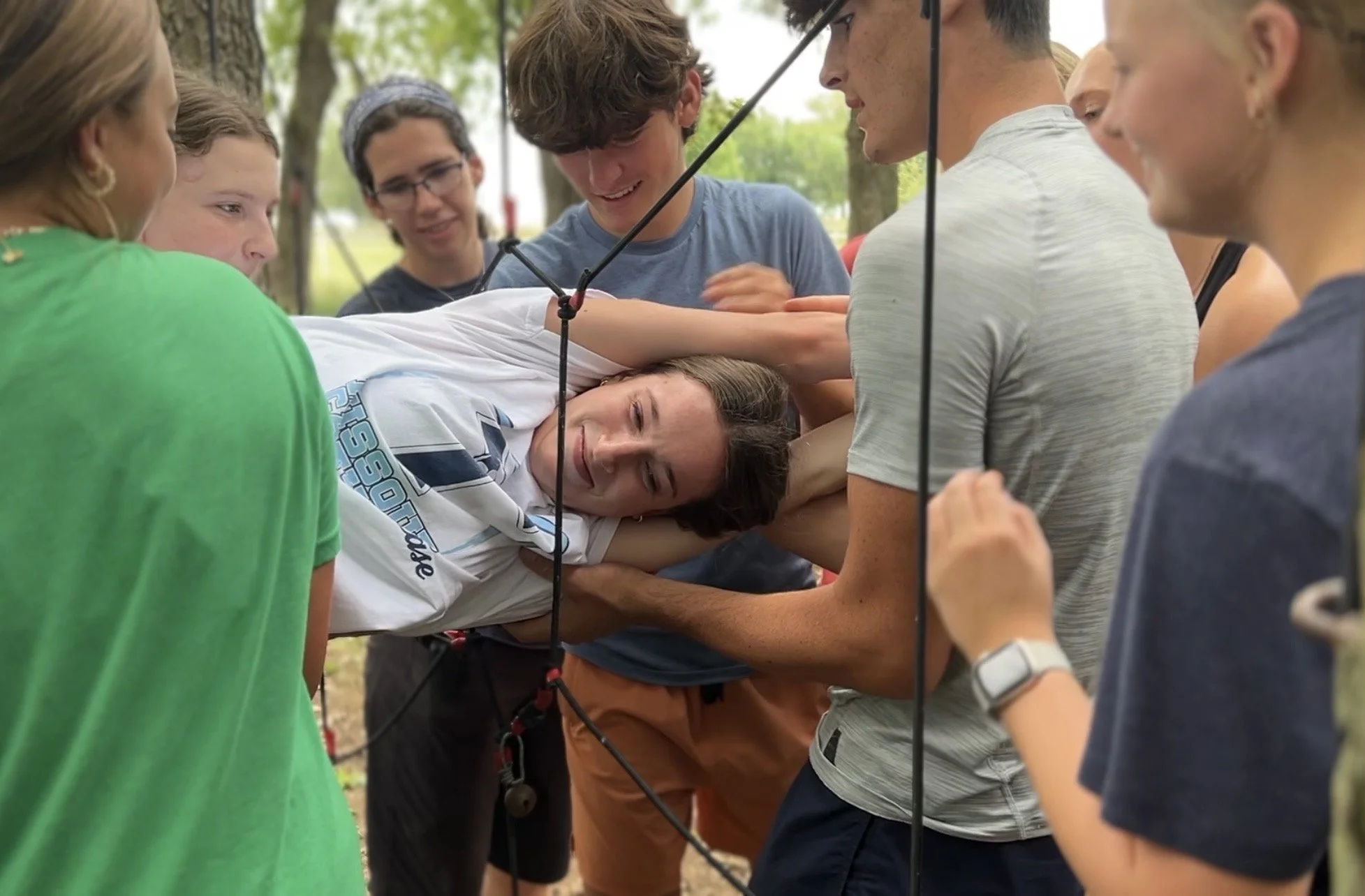 Group of young people gathered outdoors, surrounding a woman lying on the ground, smiling, as they participate in a rescue exercise involving a rescue sling.