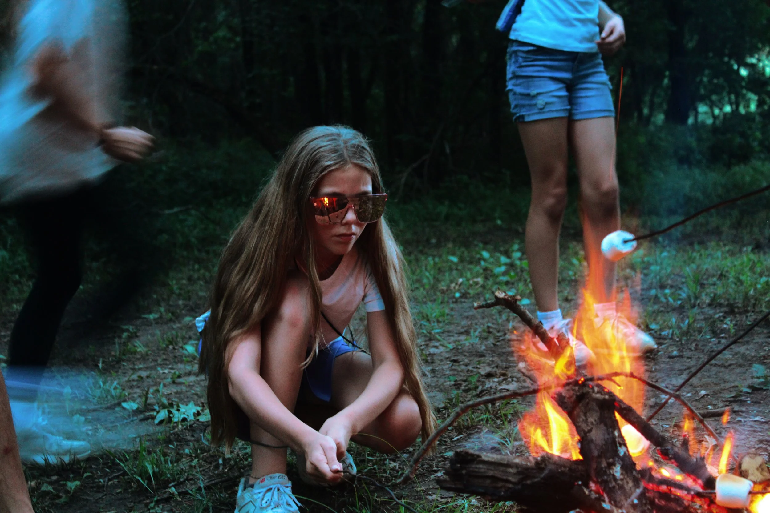 A young girl with long hair wearing sunglasses and a pink T-shirt sitting on the ground near a campfire in a forest. Other children are partially visible standing nearby.