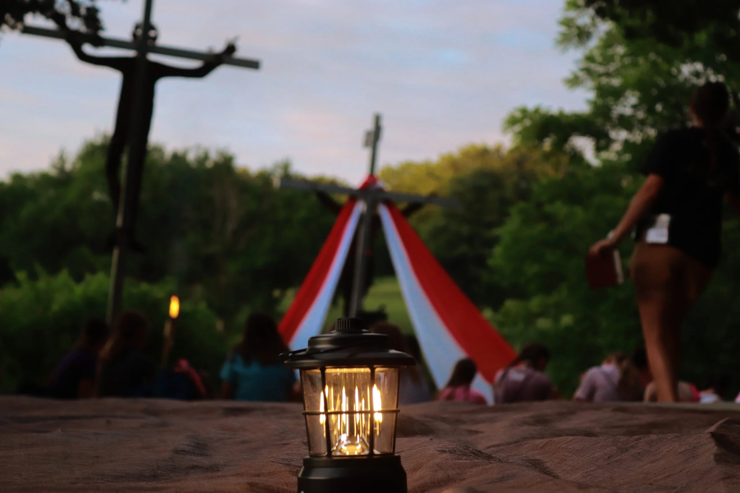 Outdoor gathering with a lit lantern on a wooden surface in the foreground, a blurred crowd seated in the background, and trees with a sky during dusk. A person is standing near a red and white tent or structure.