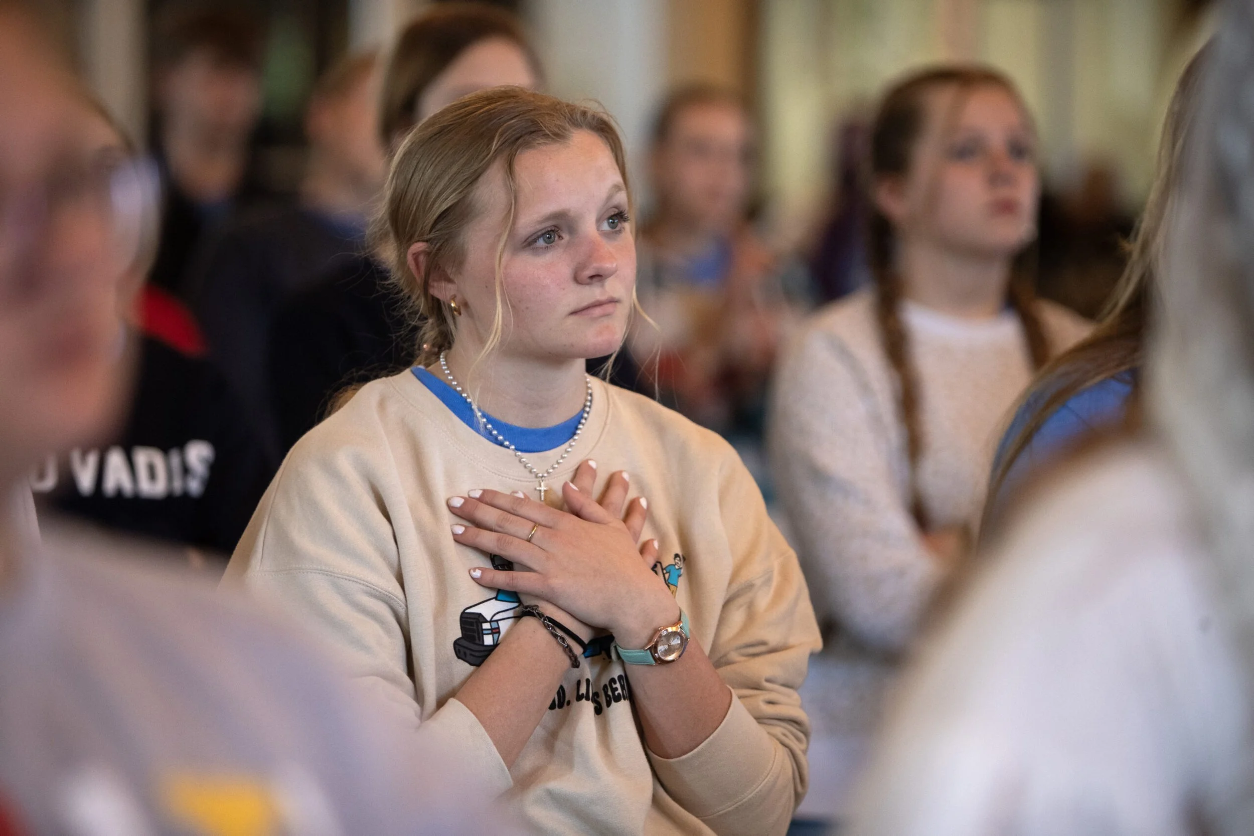 A young woman with light skin, wearing a beige sweatshirt, a pearl necklace, and various bracelets, has her hand over her heart and appears to be in a moment of reflection or prayer. She is in a room full of people, likely at an event or gathering.