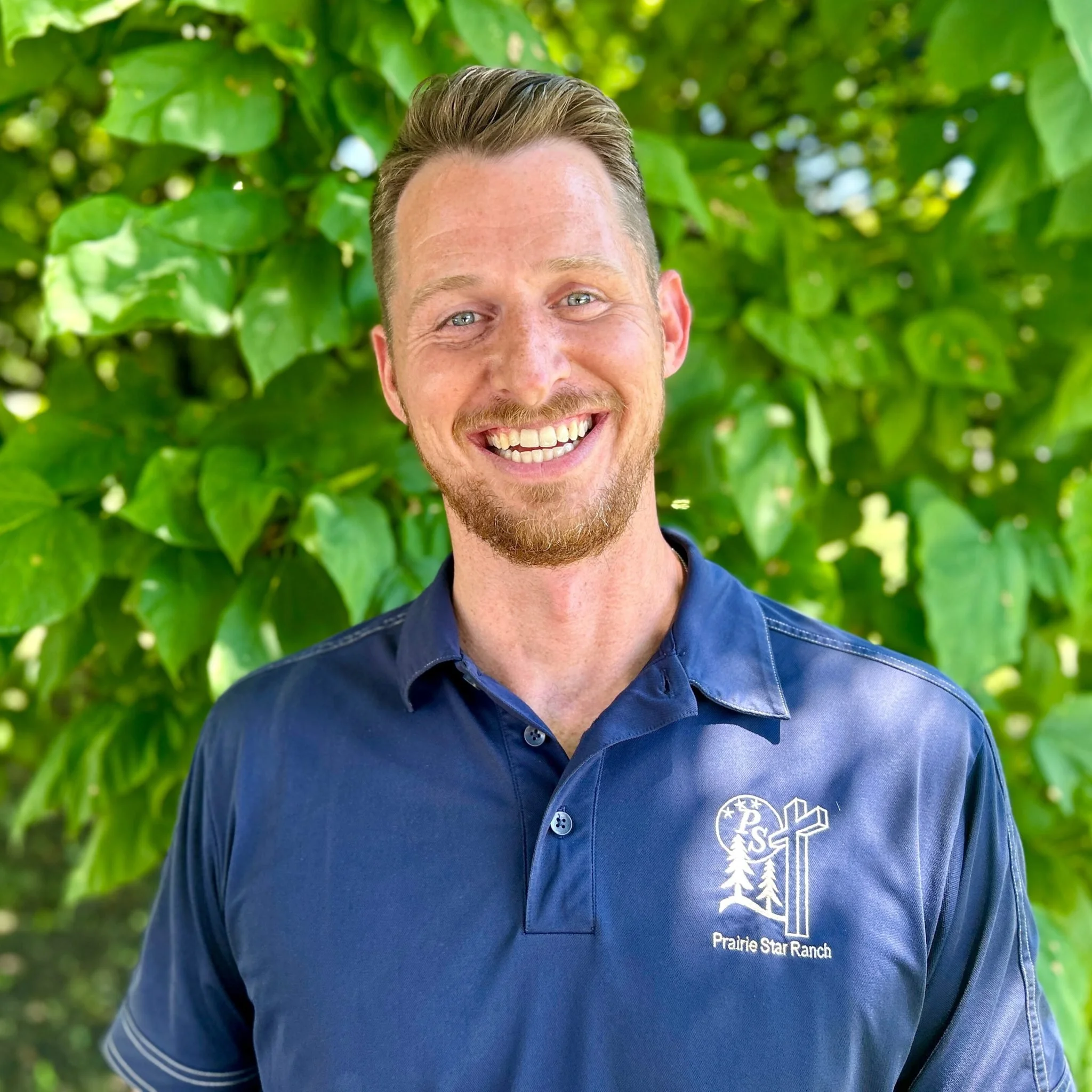 Close-up of a smiling man with short hair and a beard, wearing a navy blue shirt with a logo that says 'Prairie Star Ranch' and a tree graphic, standing in front of green foliage.