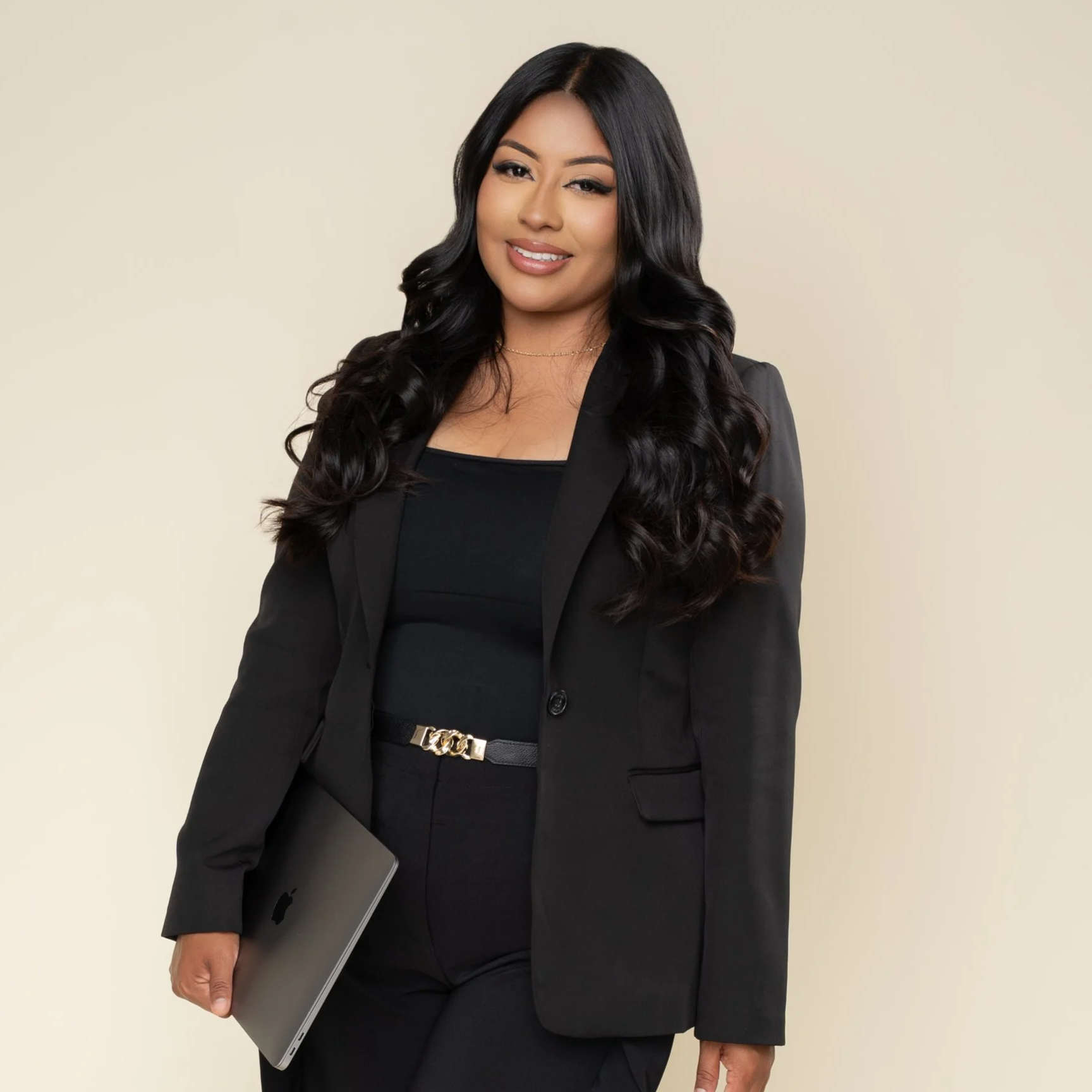 A professional woman with long black hair, dressed in a black blazer and pants, holding a silver laptop, standing against a plain light beige background.