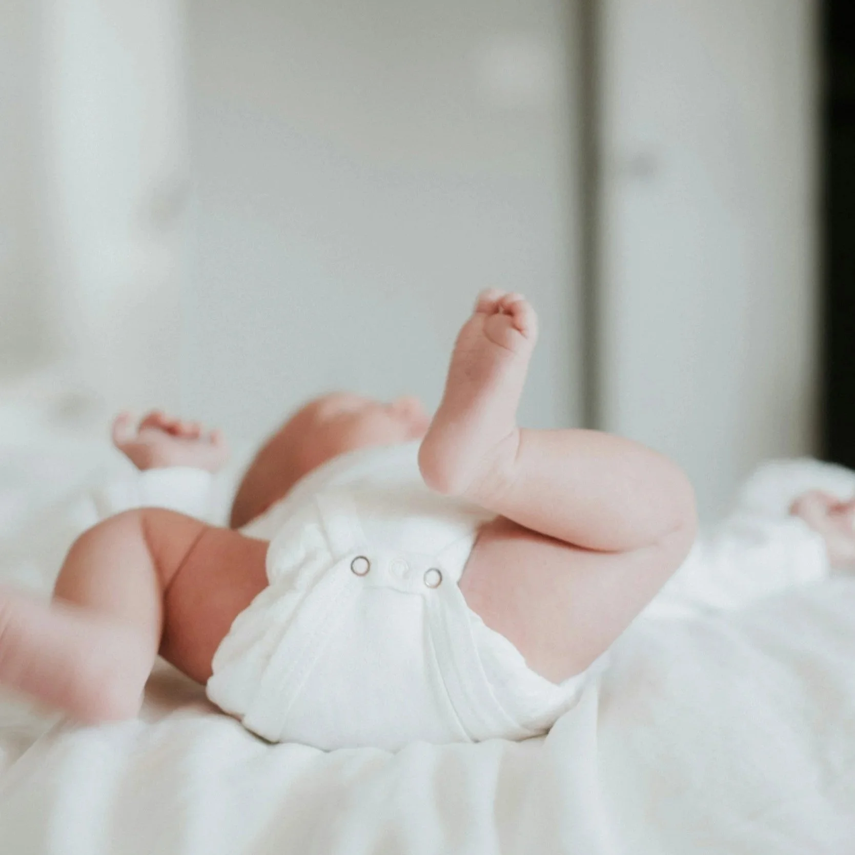 Close-up of a baby lying on their stomach on a soft surface, wearing a white onesie, with focus on the baby's legs and feet.