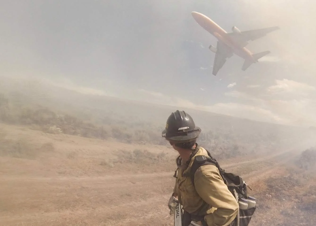 Firefighter wearing protective gear and helmet looking up at a plane flying overhead in a smoky, hazy landscape