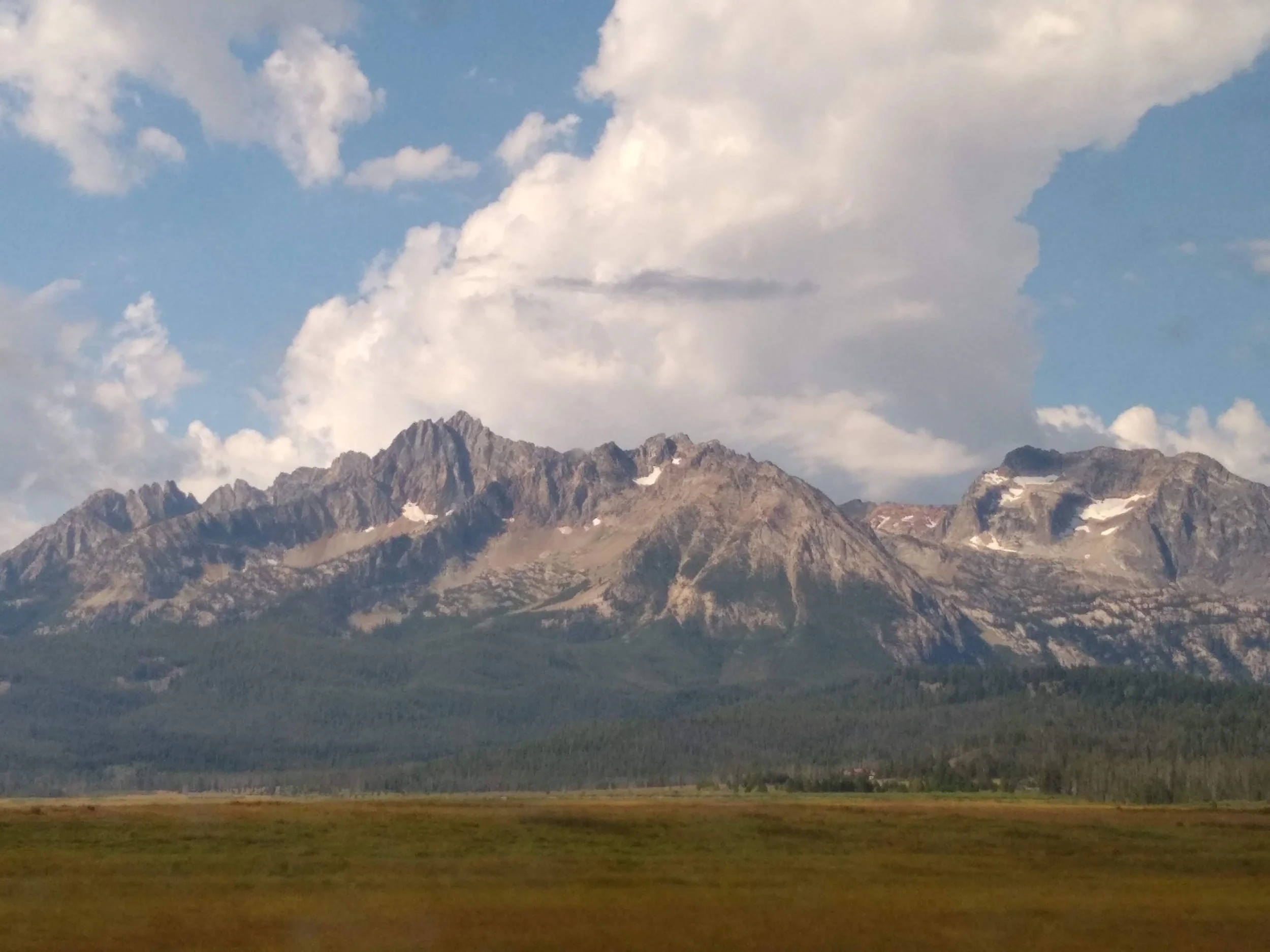 A mountain range with jagged peaks under a partly cloudy sky, with a wide green plain in the foreground.