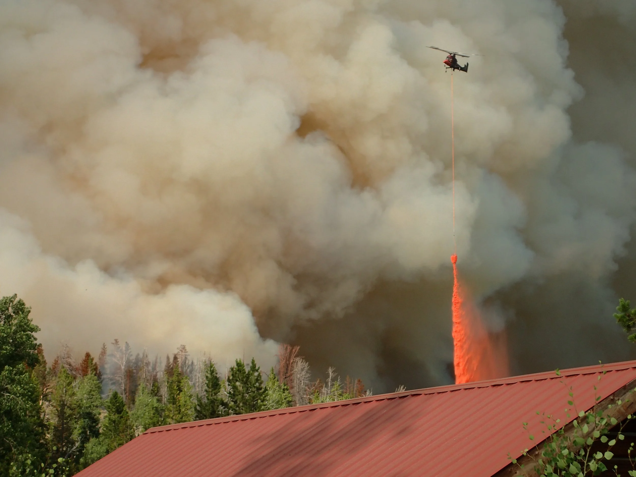 A helicopter releasing water to extinguish a wildfire in a forested area, with smoke and flames visible in the background.