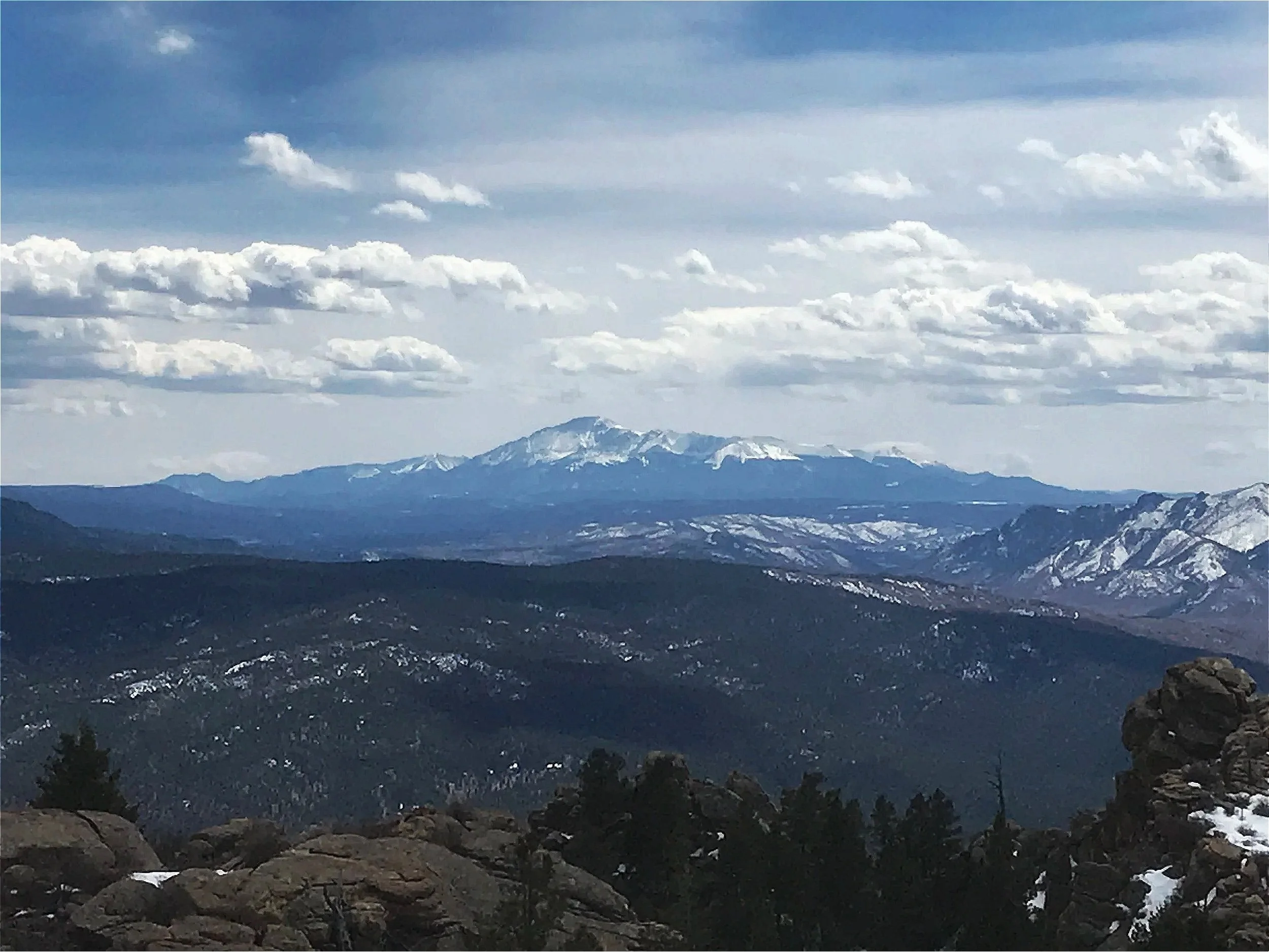 Snow-covered mountain range seen from a forested viewpoint with a cloudy sky.