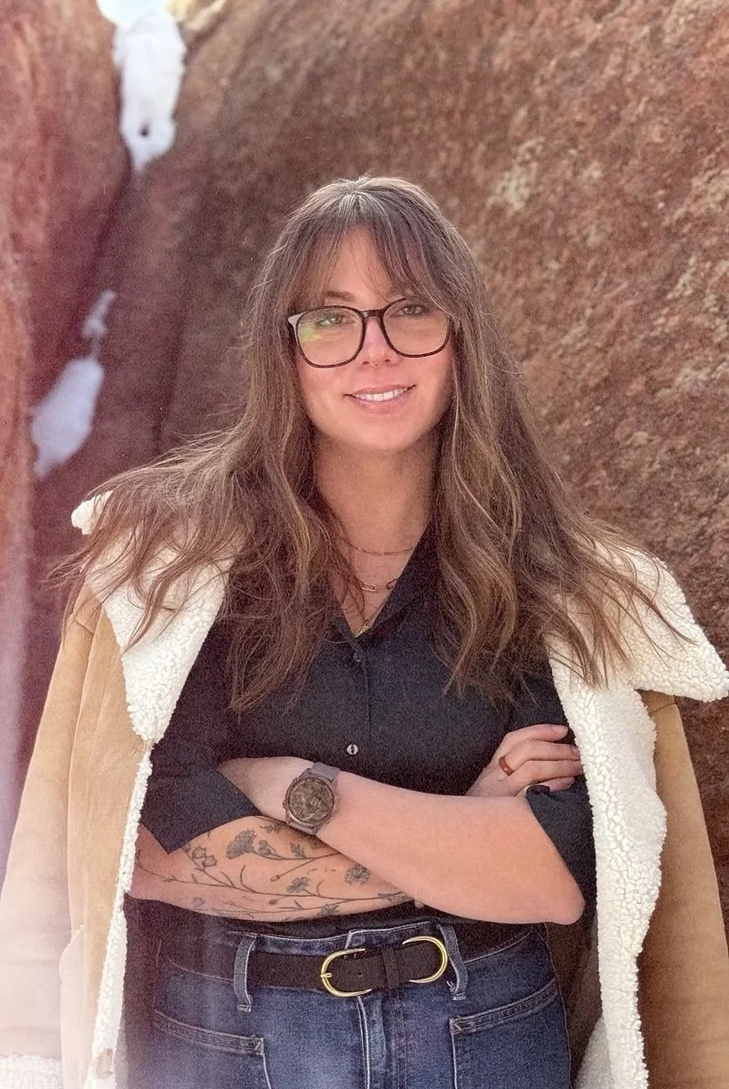 Erica Conklin with long brown hair, glasses, and tattoos on her arm, standing with her arms crossed against a rocky background, smiling at the camera.