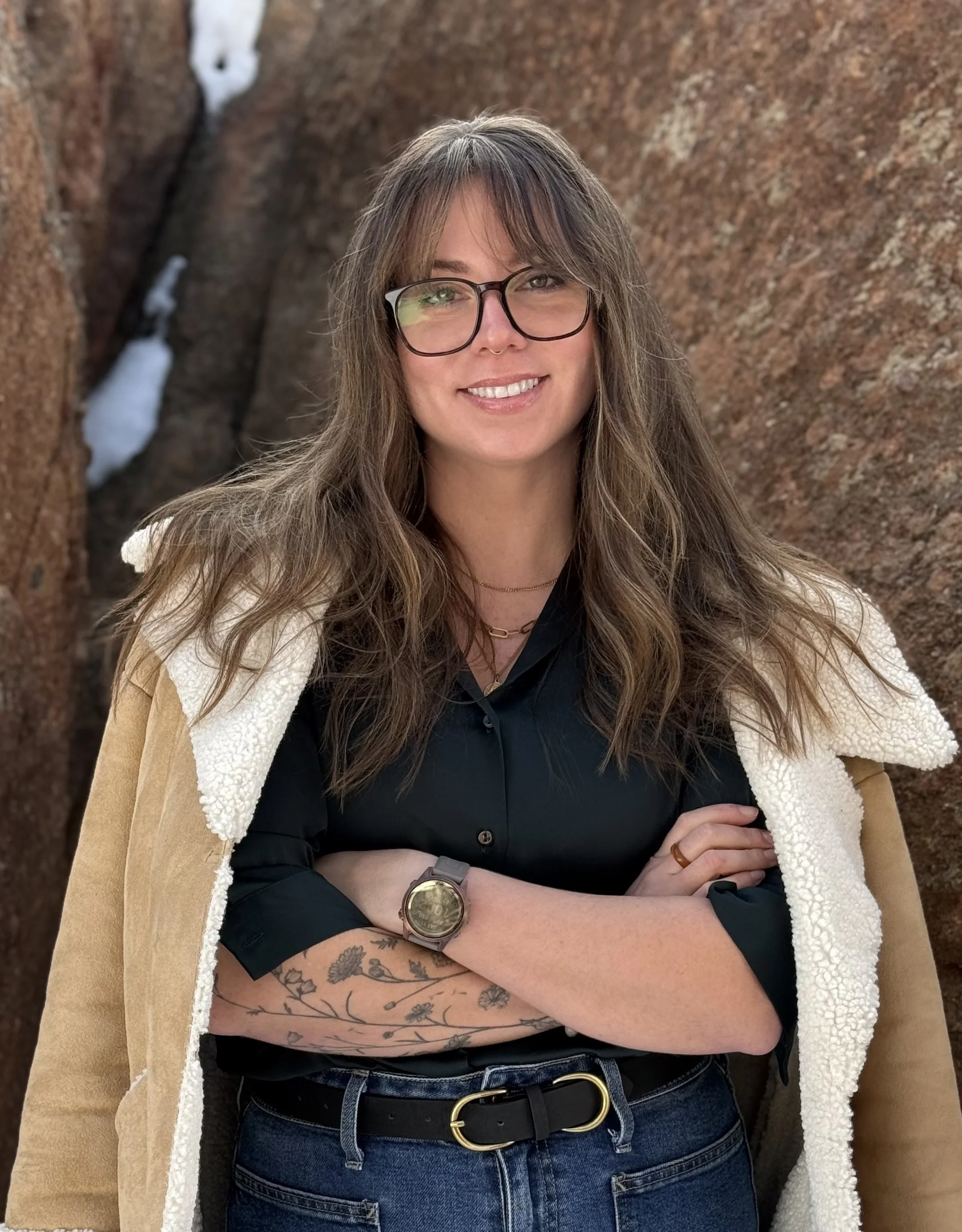 A woman with long wavy hair, glasses, and tattoos on her forearm standing outdoors against a background of large rocks with patches of snow.