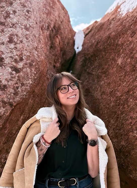 Erica Conklin with glasses and long brown hair standing between large red rock formations, smiling, with a cloudy sky visible above.