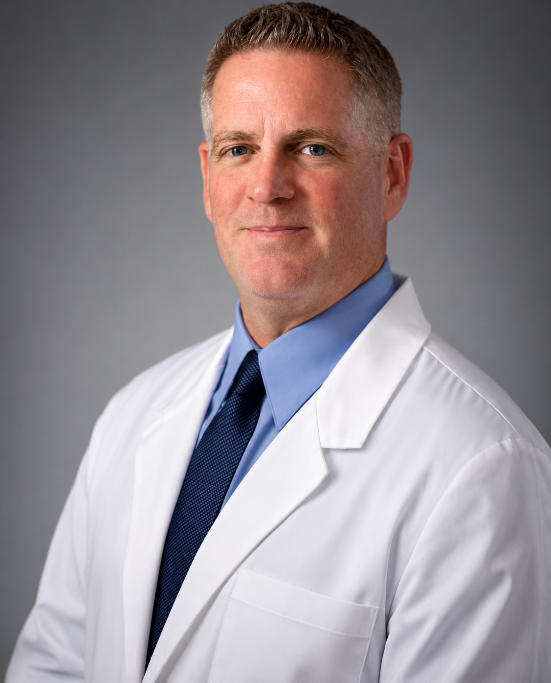 Portrait of a middle-aged male doctor wearing a white lab coat, blue shirt, and navy tie, standing against a gray background.