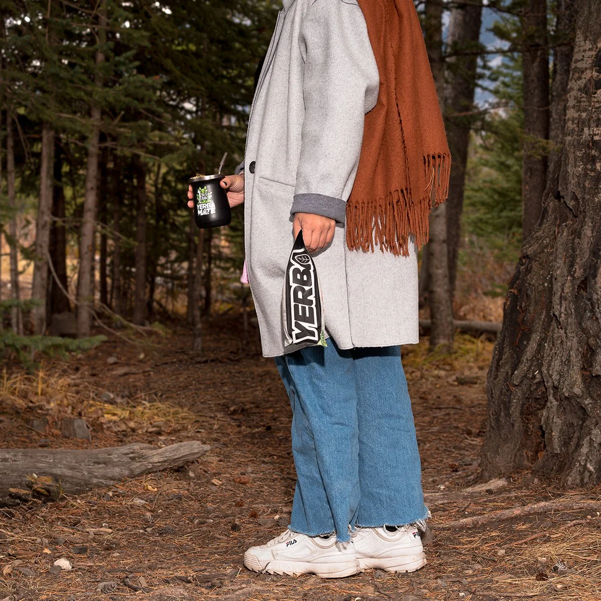 Person standing outdoors in a forest, holding a black drink tumbler and a large black and white Merrell bag, wearing a long gray coat, a brown scarf, blue jeans, and white Fila sneakers.
