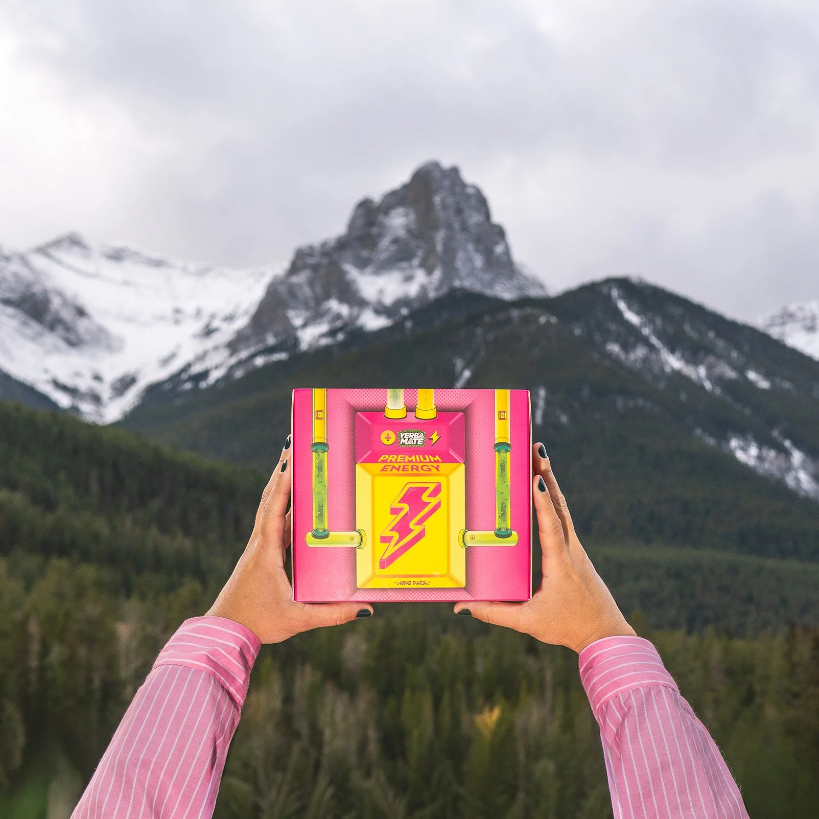 Person holding a colorful energy drink box with mountains in the background.