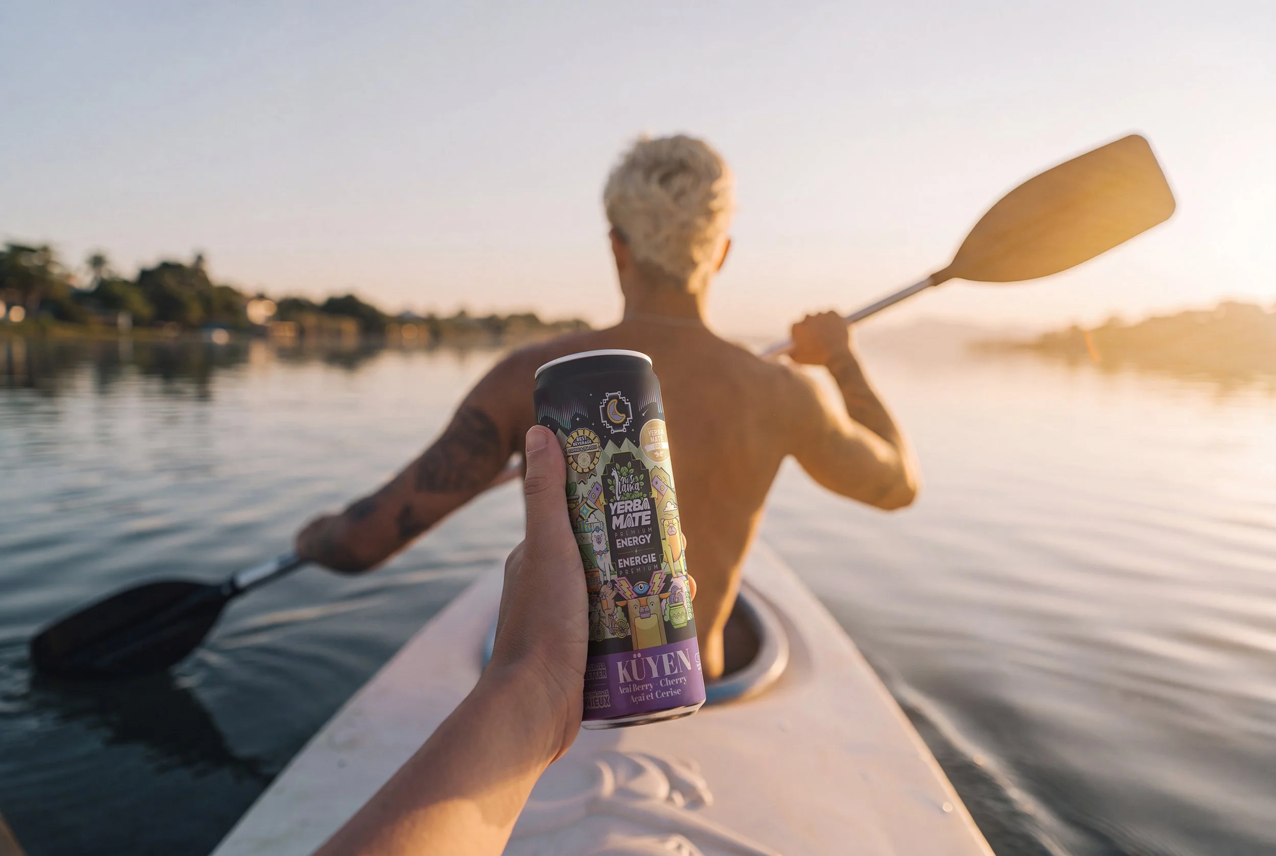 Person holding a can of energy drink while kayaking on a river during sunset, with a man paddling in the background.
