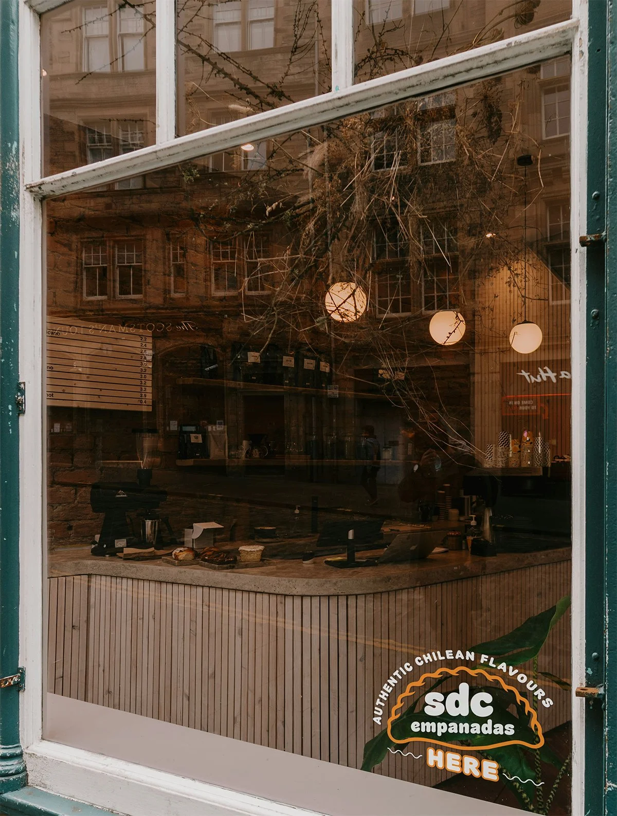 View of a bakery or cafe through a window with a sign that reads 'sdc empanadas here', showing a counter with baked goods and warm lighting inside.