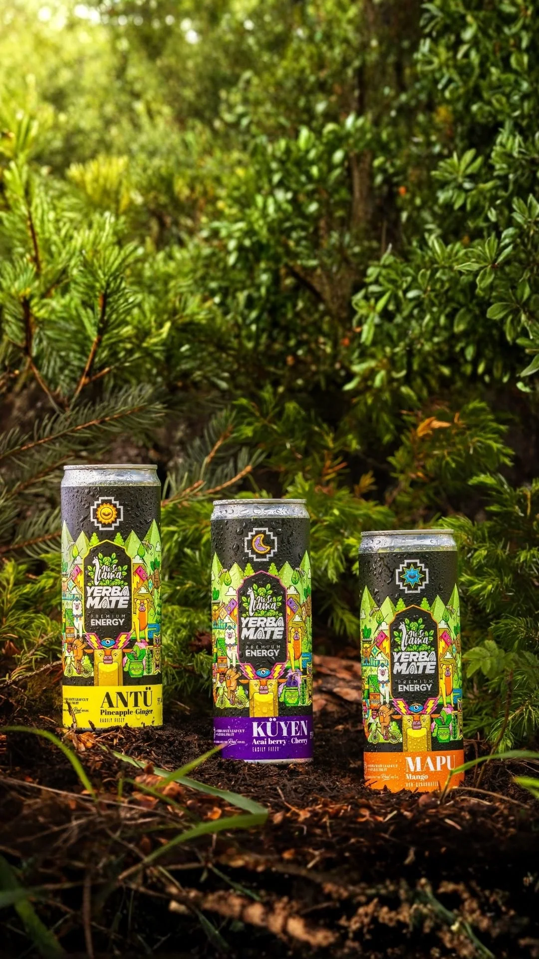 Three cans of energy drinks with colorful, nature-themed labels placed on the ground in a forest setting with green bushes in the background.