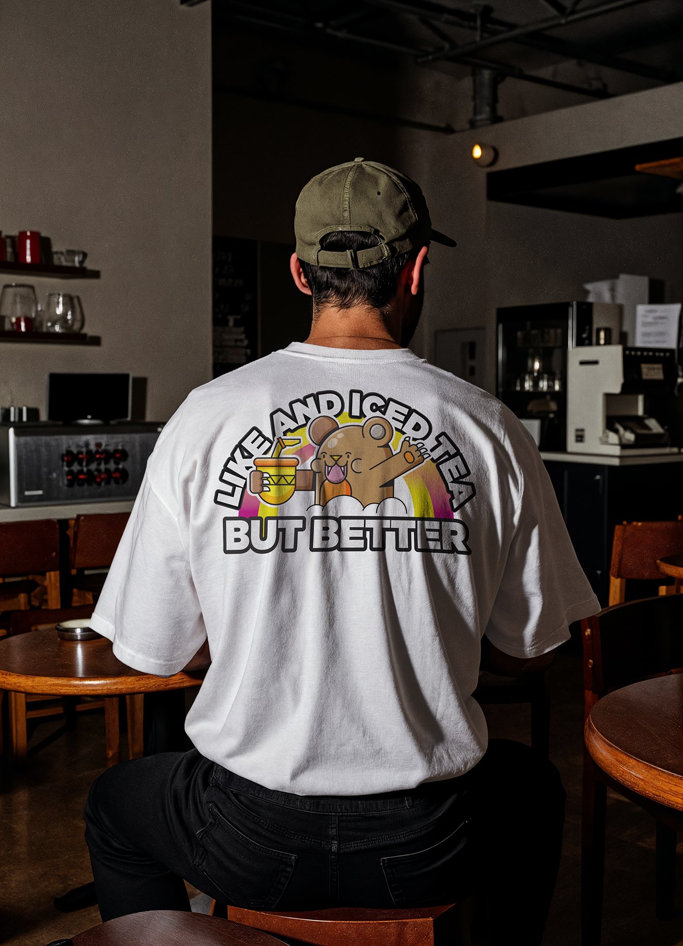 Back view of a person wearing a white t-shirt with a design featuring a cute bear holding a drink and the text "LIKE AND I GET TREATED BUT BETTER". The individual is sitting in a cafe with wooden chairs and tables, and a coffee shop interior in the background.