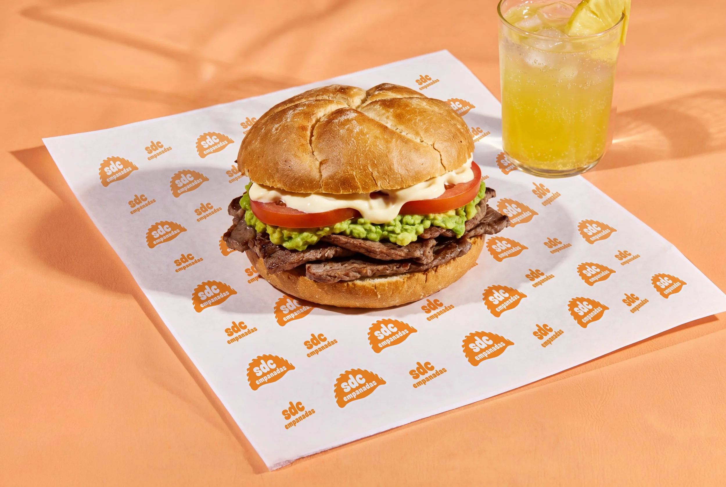 A sandwich with beef, guacamole, tomato, mayonnaise, and a sandwich bun on branded paper, next to a glass of lemon soda with ice.