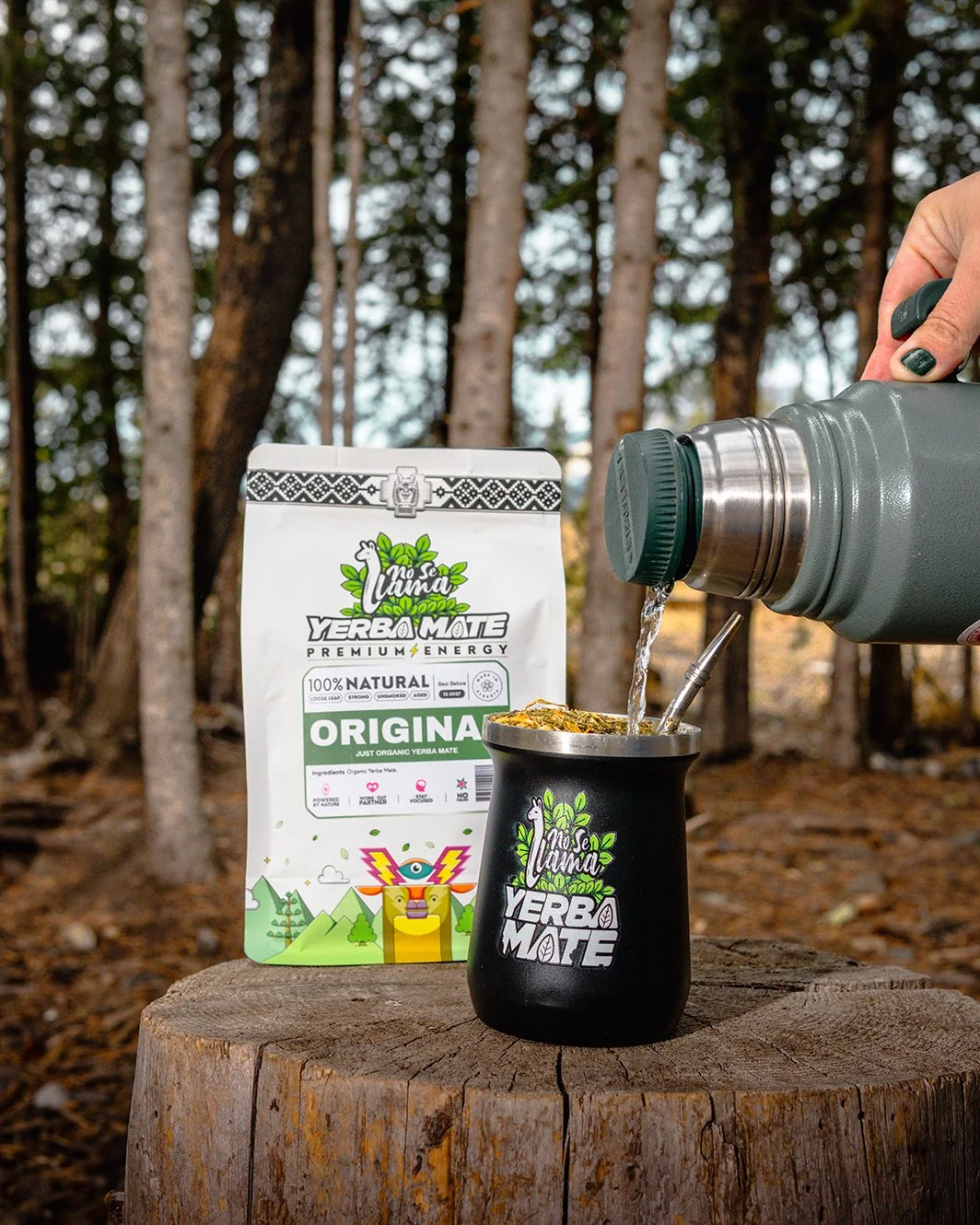 Yerba Mate tea being poured from a thermos into a black wooden mate cup with the 'No Se Toma Yerba Mote' logo, beside a package of Yerba Mate on a tree stump in a forest background.