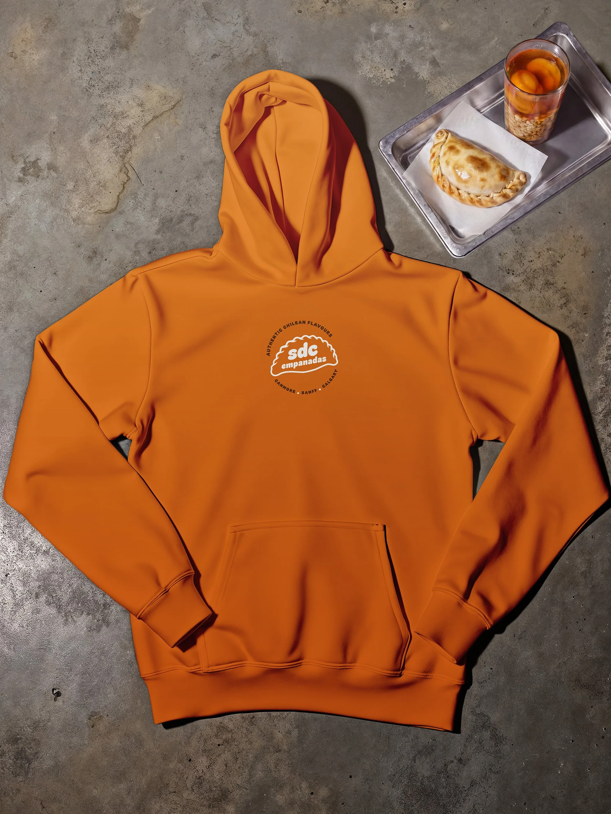 An orange hoodie with a logo on the front reading 'sdC empanadas' resting on a concrete floor next to a metal tray with a baked empanada and a glass of tea with fruit.
